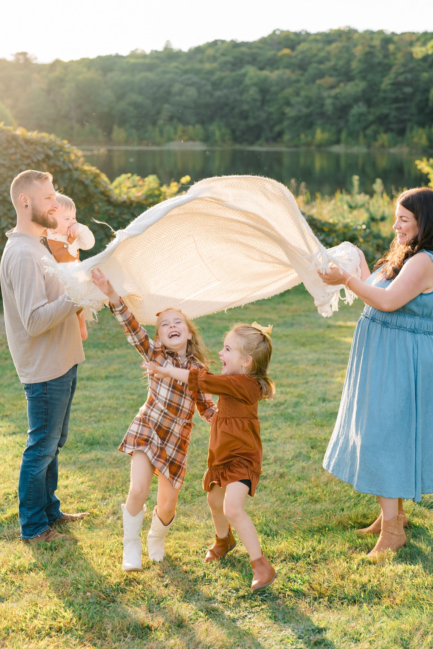 A joyful family plays with a blanket outdoors at golden hour near a lake, with two girls laughing in fall outfits.