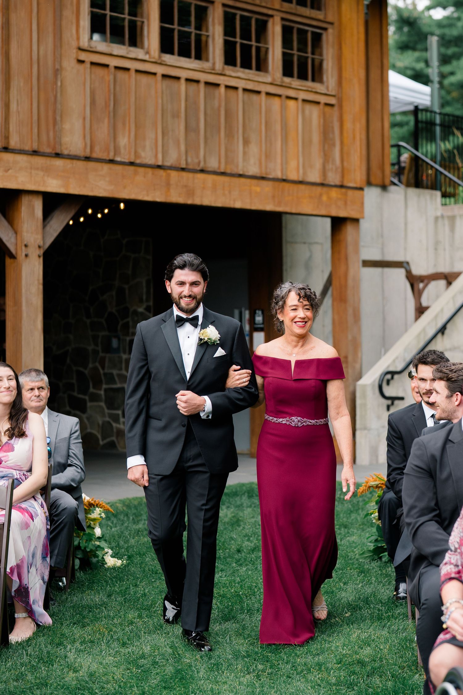 Groom in black tuxedo walks down wedding aisle with woman in burgundy off-shoulder gown at outdoor barn venue.