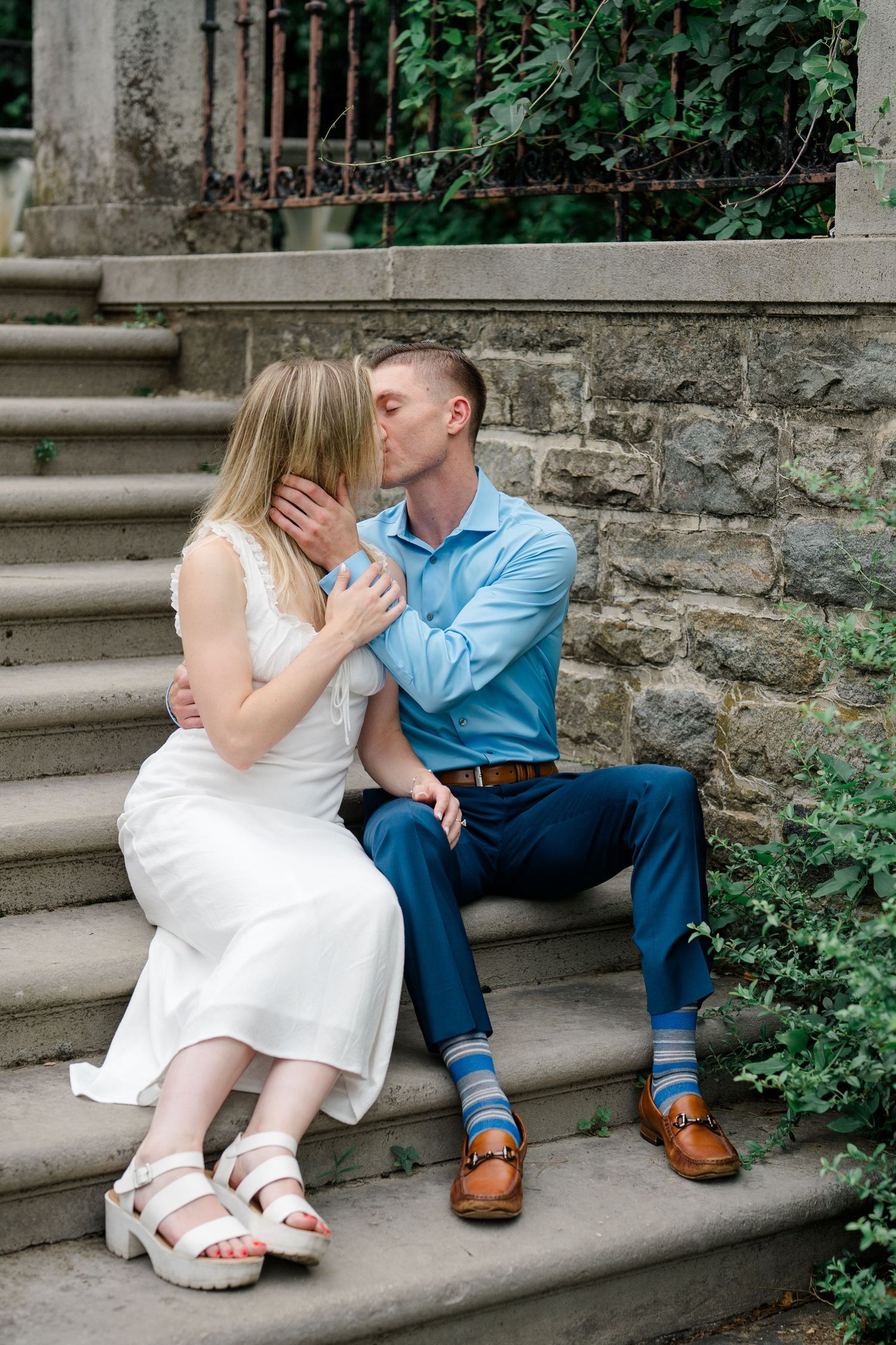 Engaged couple sharing a romantic kiss while seated on stone steps surrounded by lush greenery.