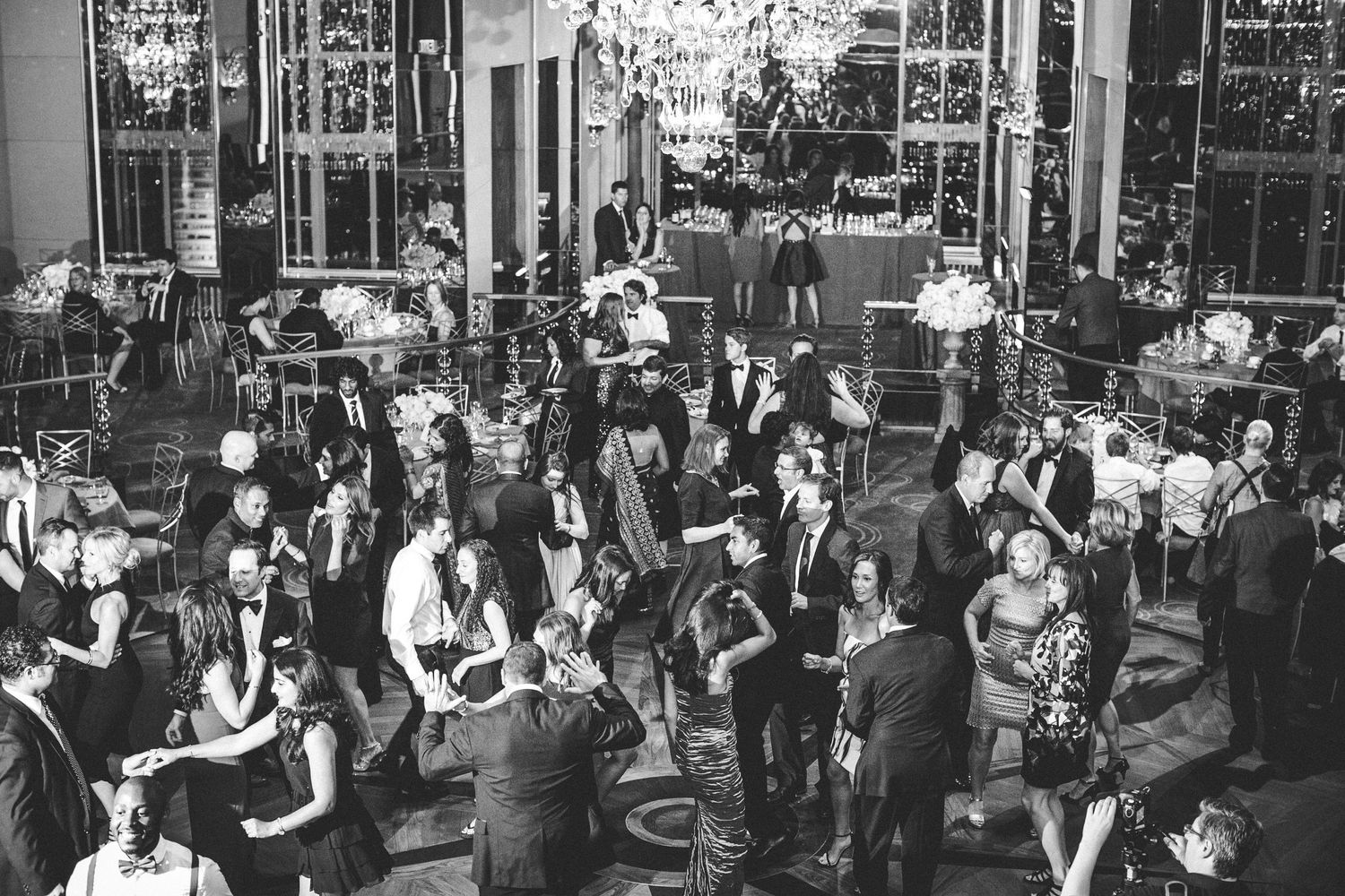 A black and white photo of a crowded wedding reception with guests dancing and socializing under elegant chandeliers.