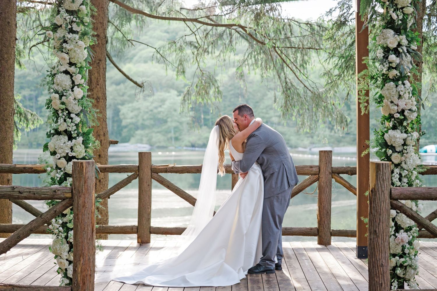 Romantic moment between newlyweds on a flower-adorned wooden deck with forest and lake views.
