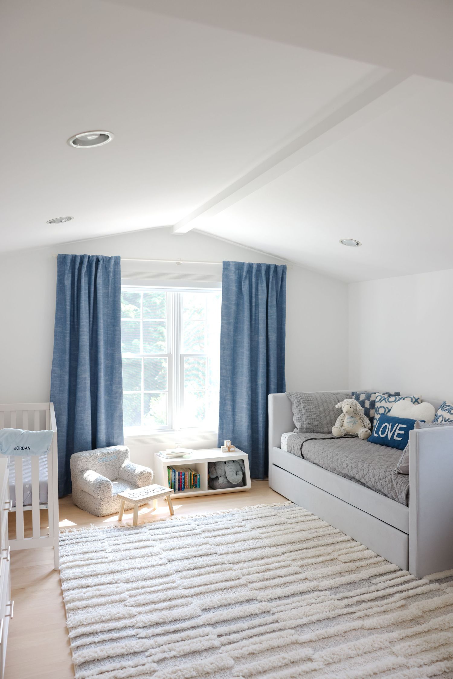 A cozy nursery room with blue curtains, gray daybed, striped rug and white crib under sloped ceiling.