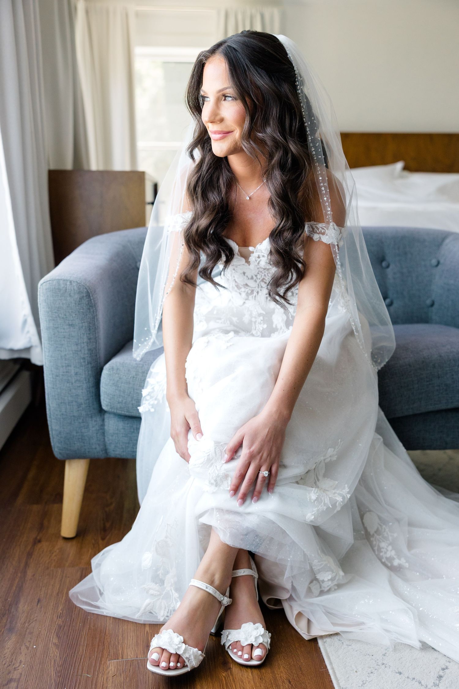 A bride in a white lace gown sits on a blue chair showing off her sparkly wedding shoes.