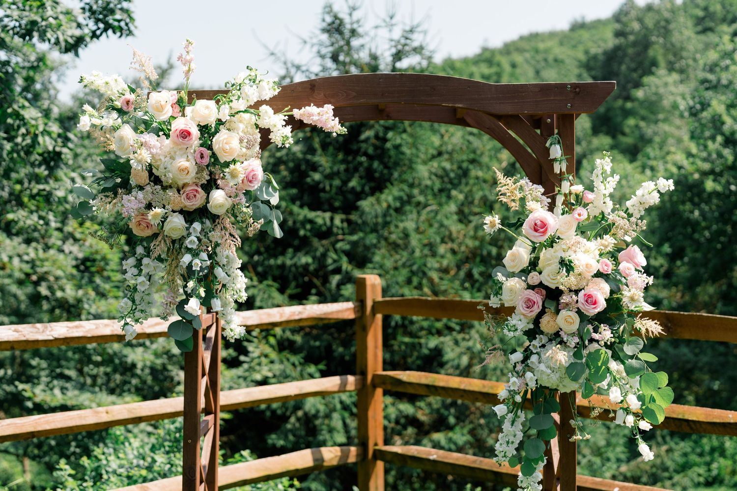Wooden wedding arch decorated with lush pink and white floral arrangements in outdoor setting.