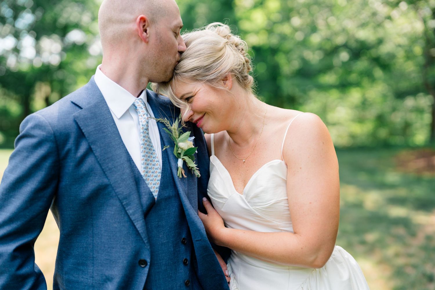 Bride and groom share a tender moment outdoors, groom kissing bride's forehead in wedding attire.