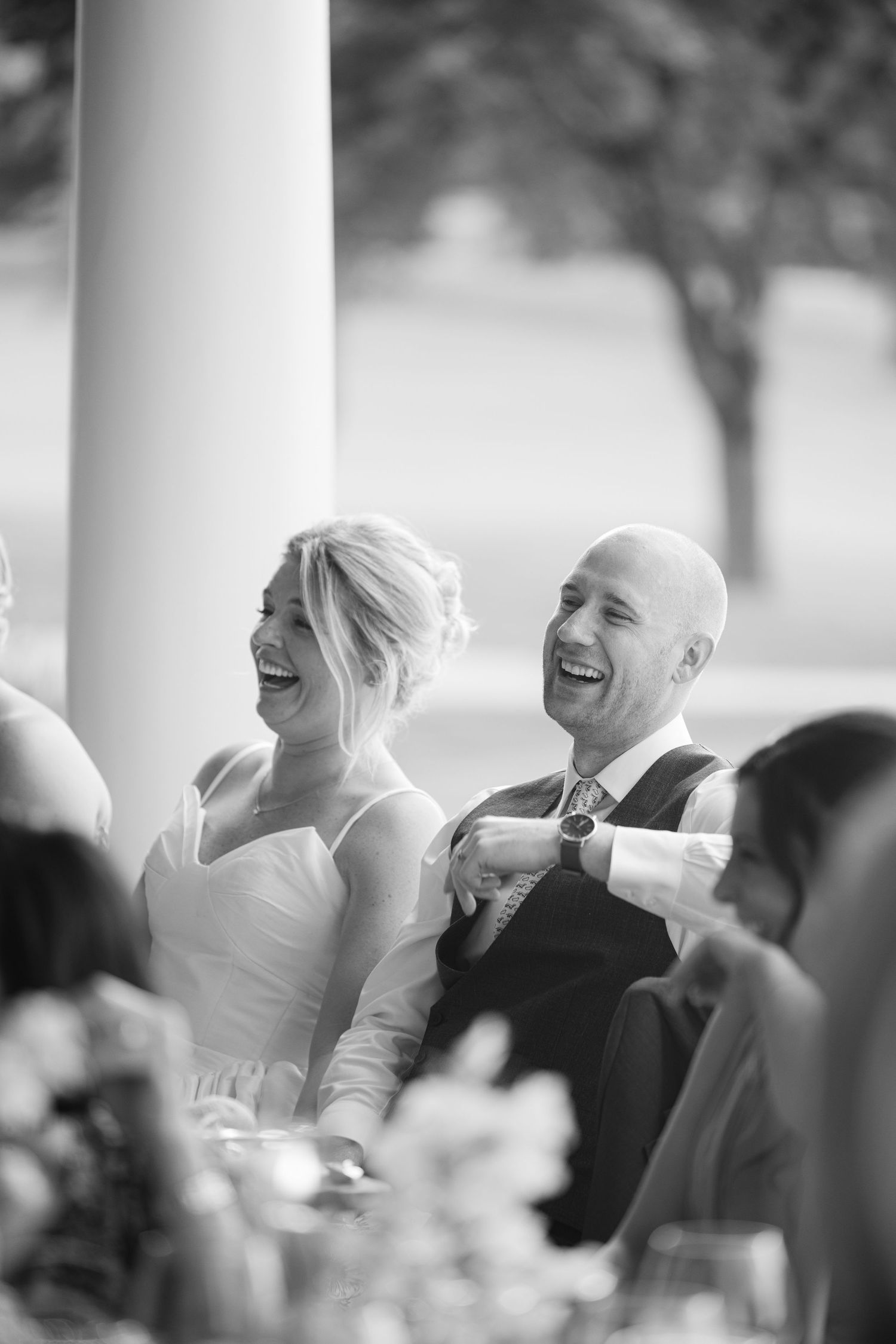 Black and white photo of a joyfully laughing bride and bald groom seated together during their wedding reception.