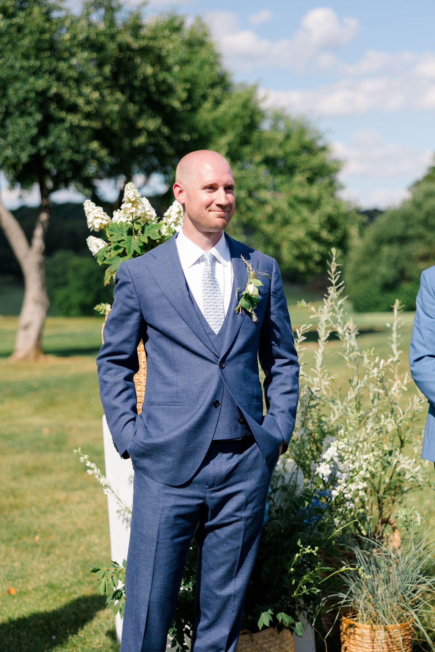 Groom in navy blue three-piece suit smiling outdoors at garden wedding ceremony with floral arrangements.