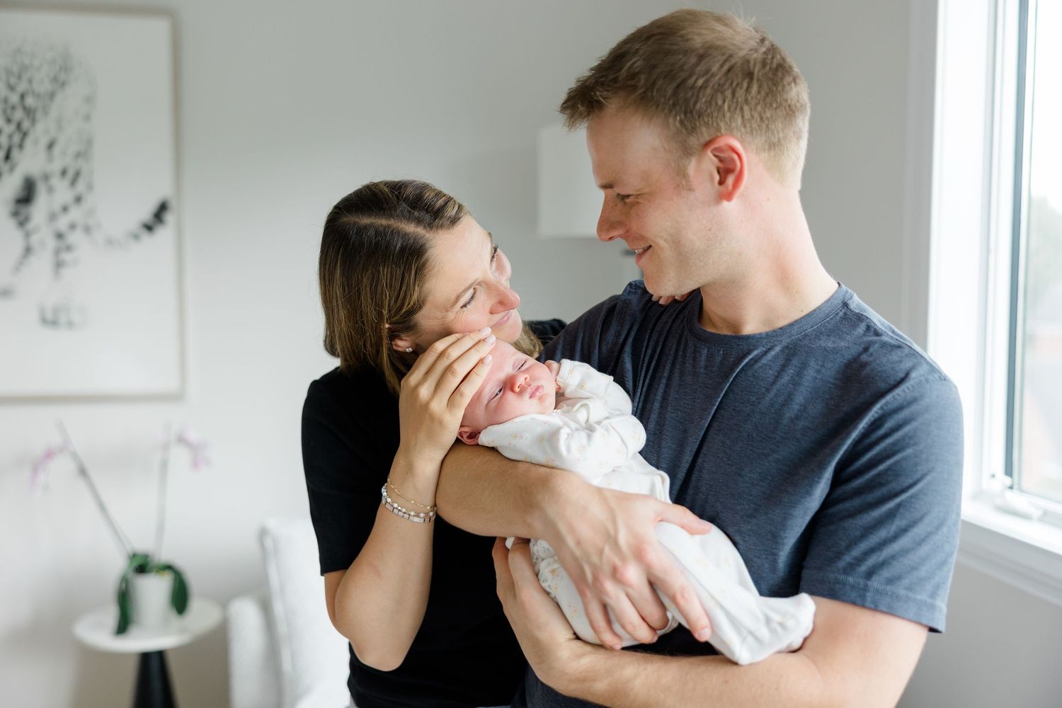 A parent in a navy shirt holds and tenderly comforts a swaddled baby near a bright window.