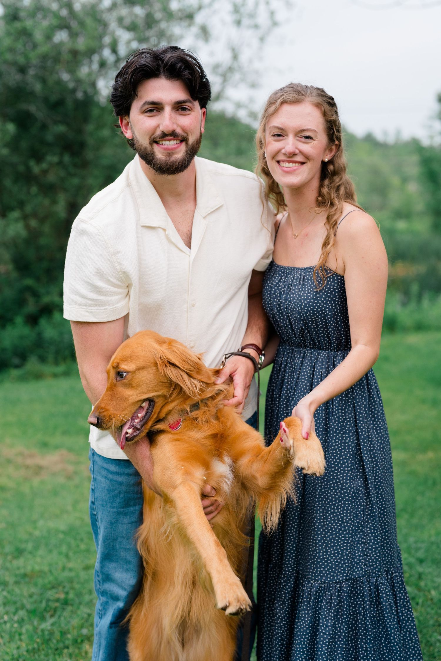 A couple in formal attire stands outdoors with their playful golden retriever dog on grassy field during photoshoot.
