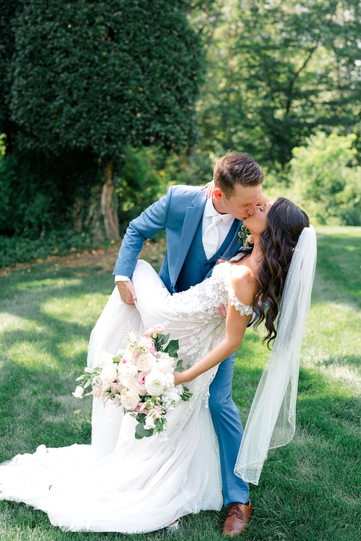 Newlyweds embrace on grassy lawn with bride's white dress flowing and groom in bright blue suit.