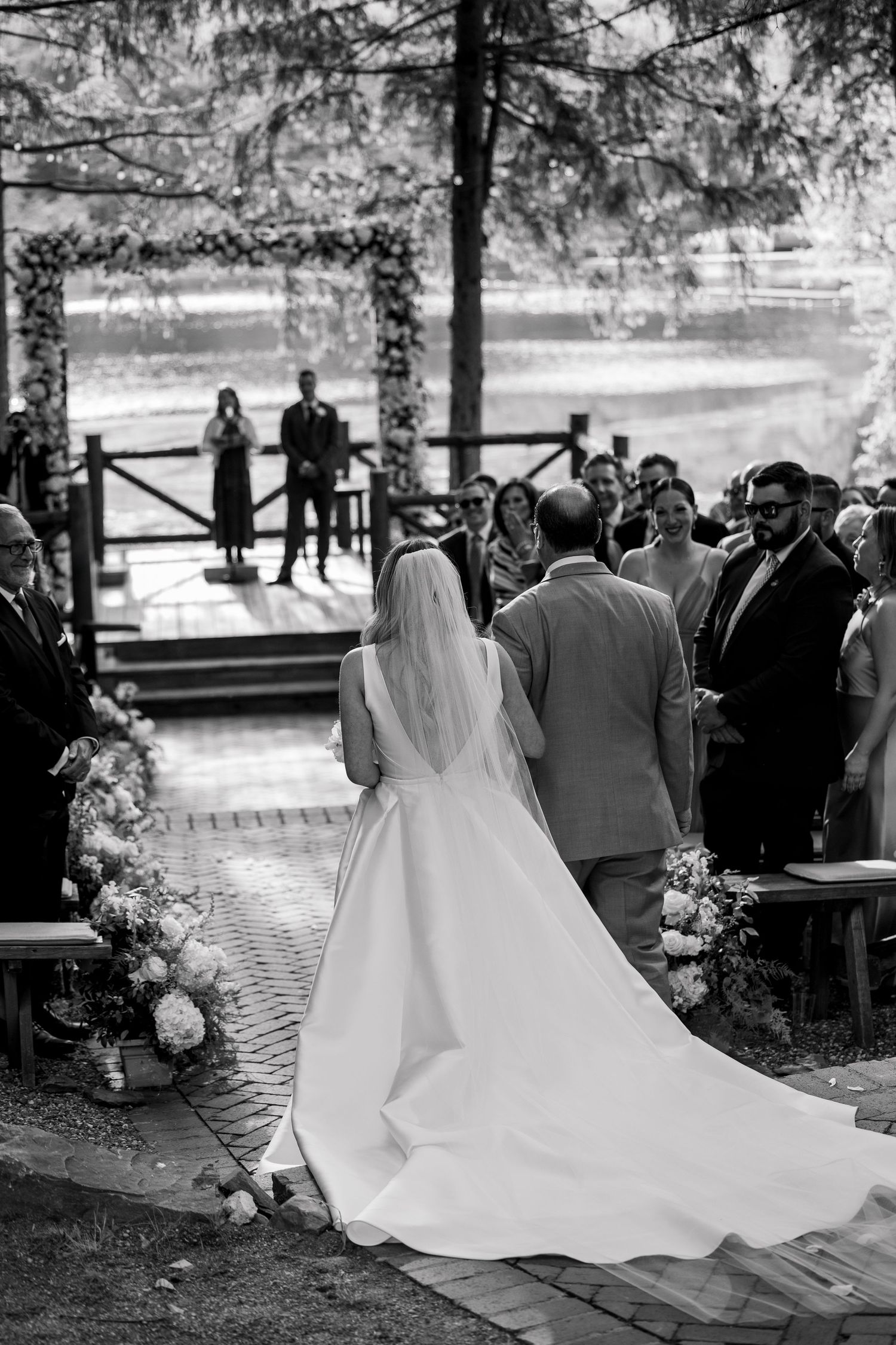 Black and white wedding ceremony by a lake with guests watching beneath tall pine trees.