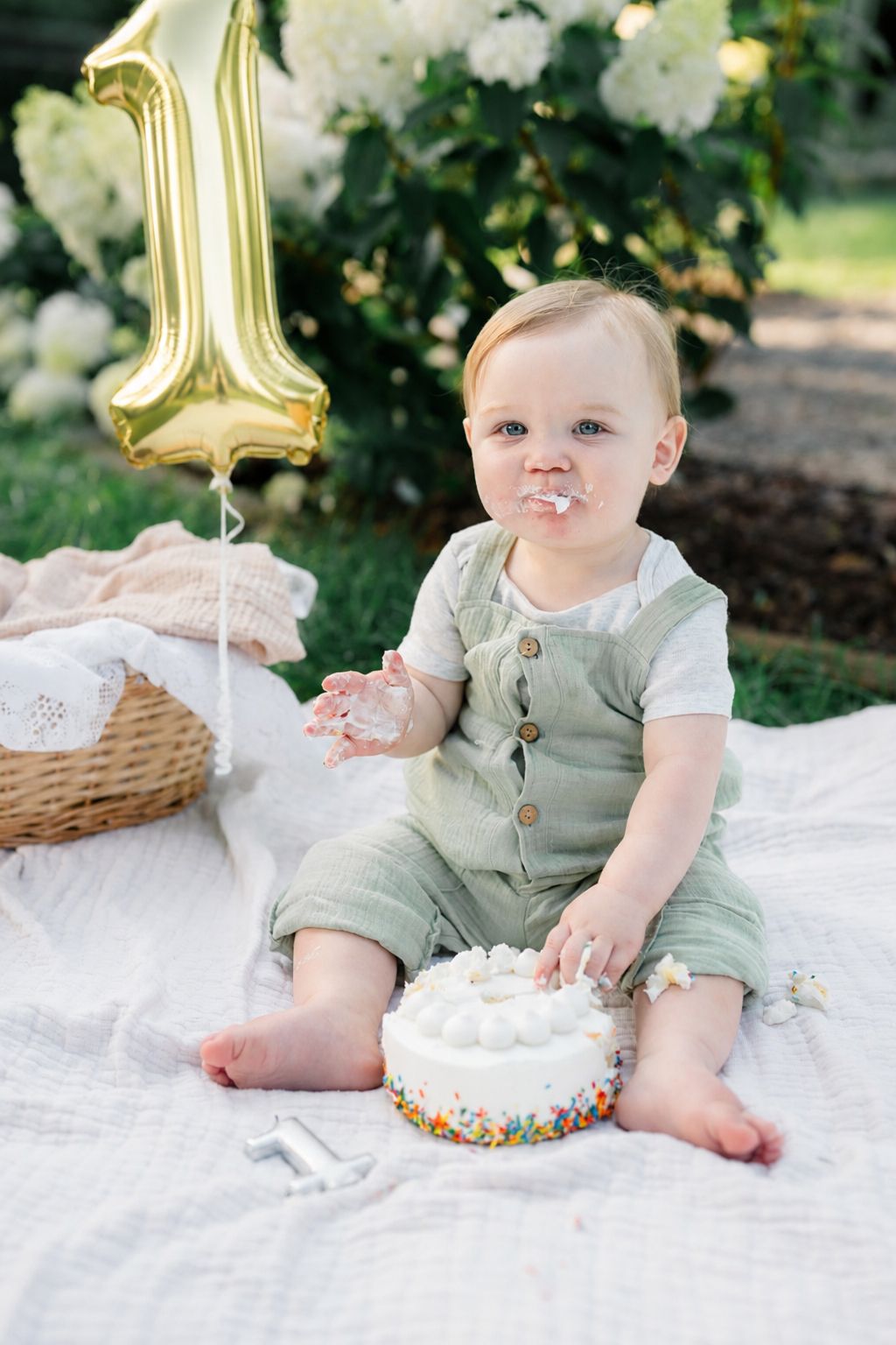 Baby celebrates first birthday outdoors, smashing white cake with sprinkles, gold number one balloon nearby.