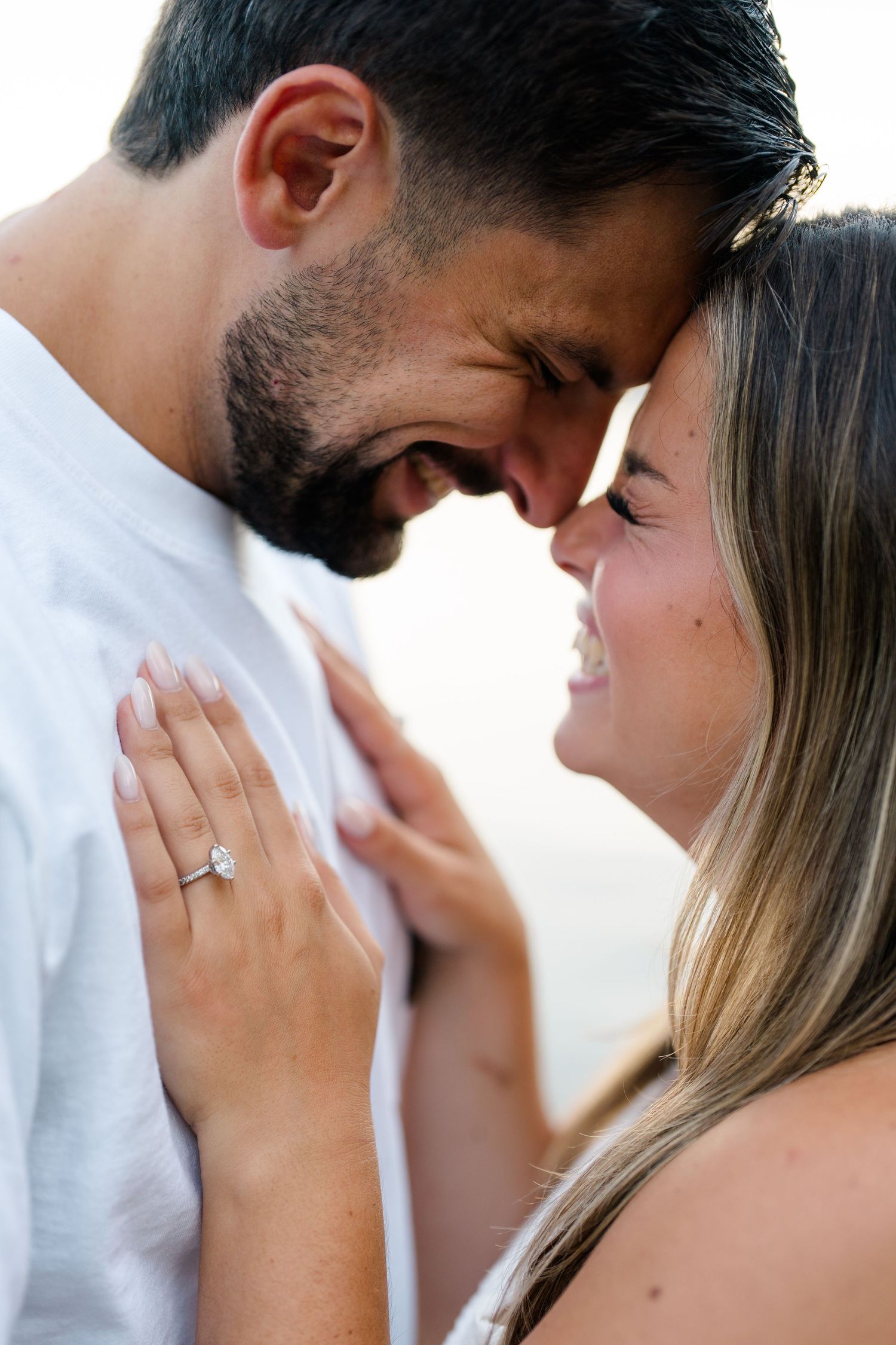 Happy couple sharing a romantic moment, woman wearing diamond engagement ring, foreheads touching and smiling.