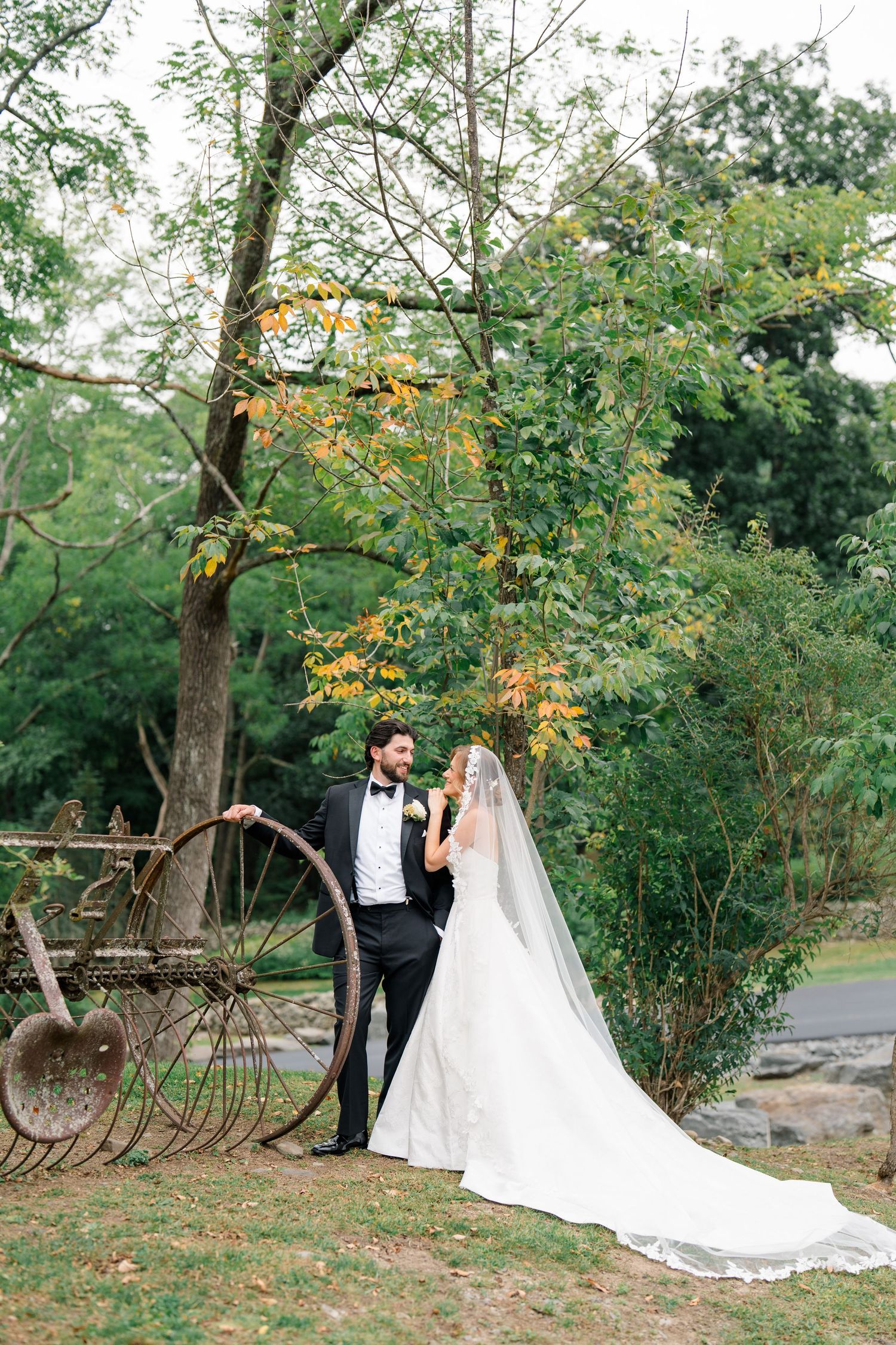 Bride and groom pose near antique farm equipment under autumn trees, bride wearing long lace veil and elegant gown.