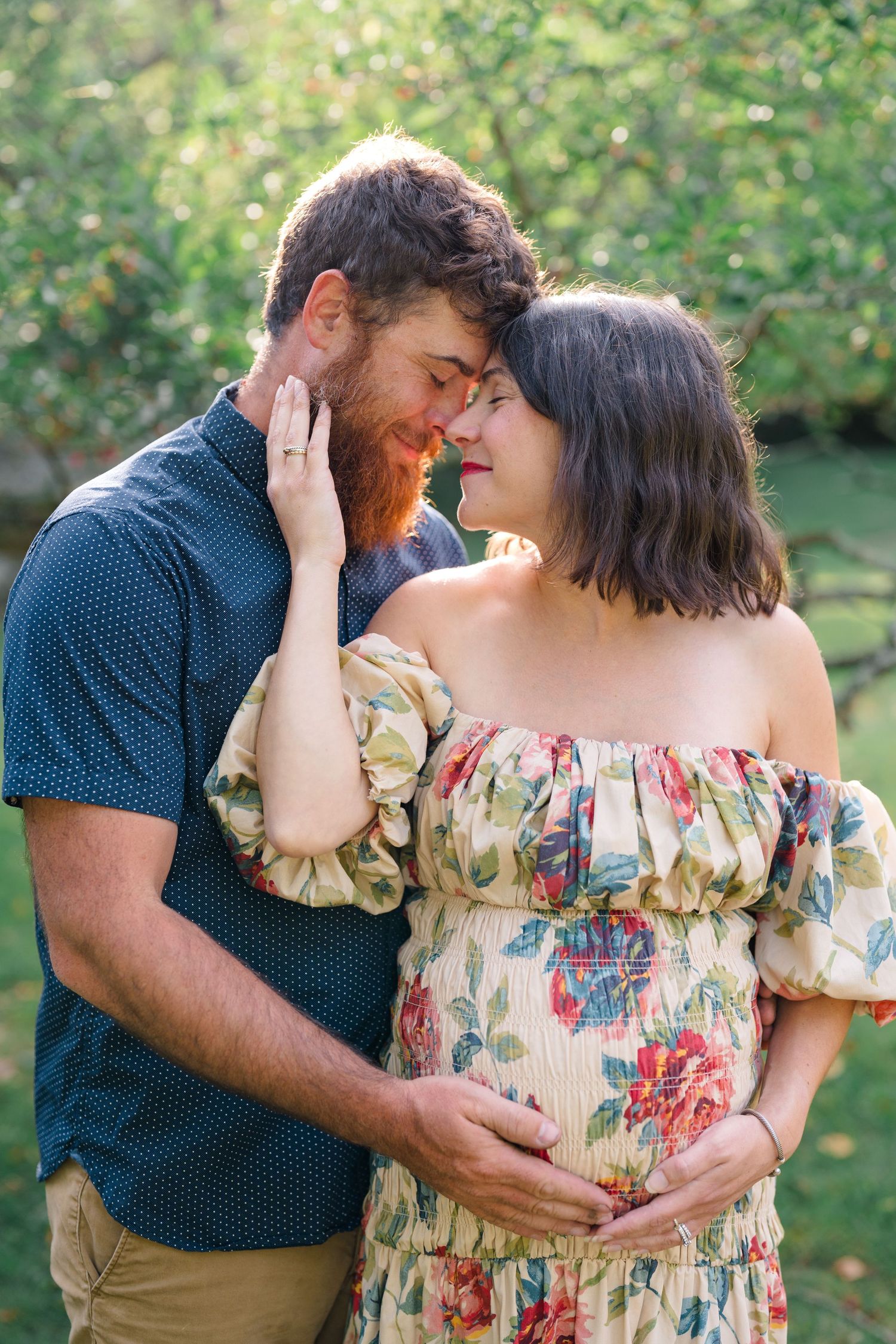 Pregnant couple sharing a tender moment outdoors, woman in floral off-shoulder dress, man with red beard gently touching her face.
