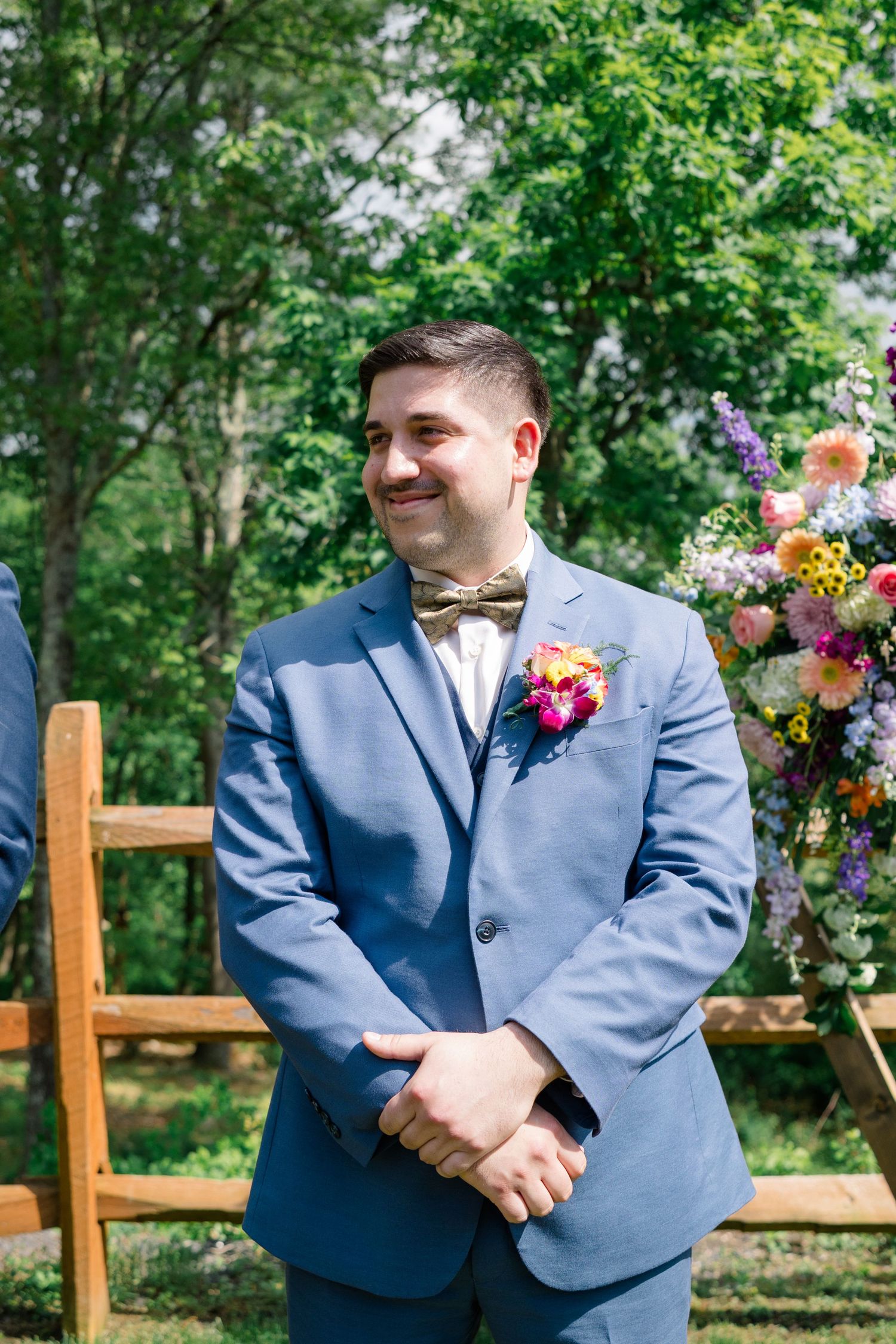 Groom in light blue suit with bow tie and boutonniere smiles during outdoor wedding ceremony.