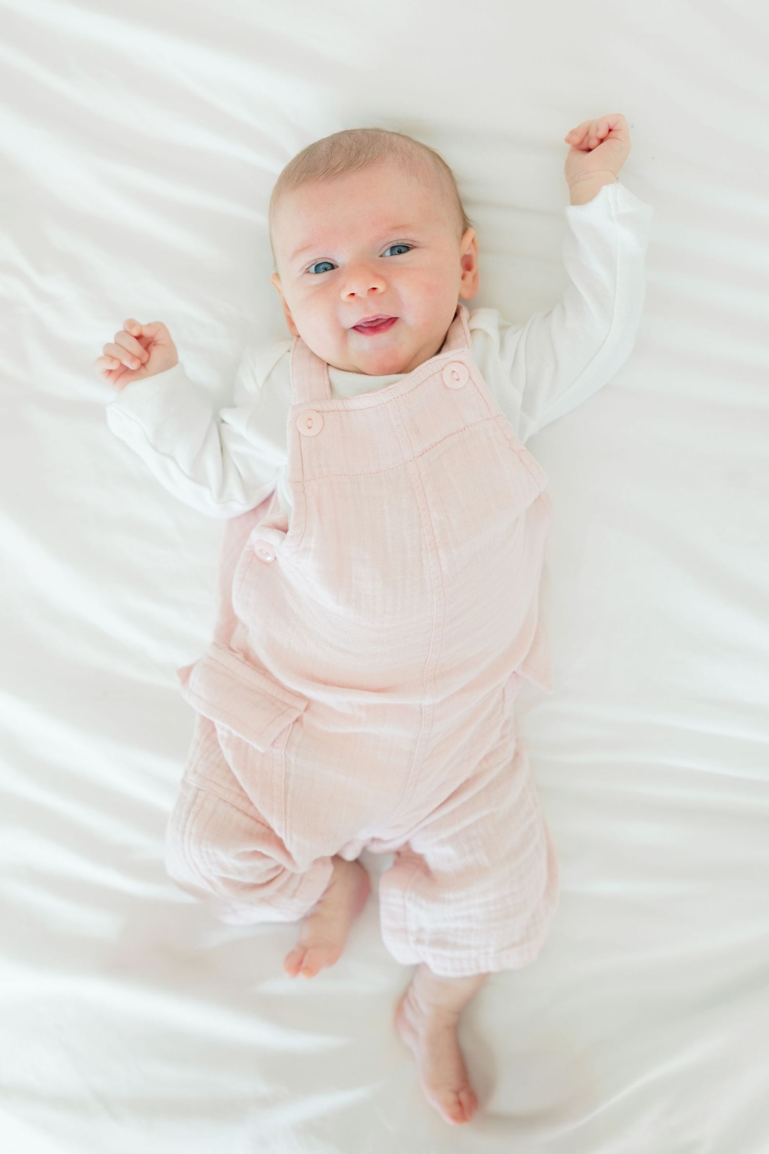 Baby in soft pink outfit lying on white bedding while making adorable expressions.