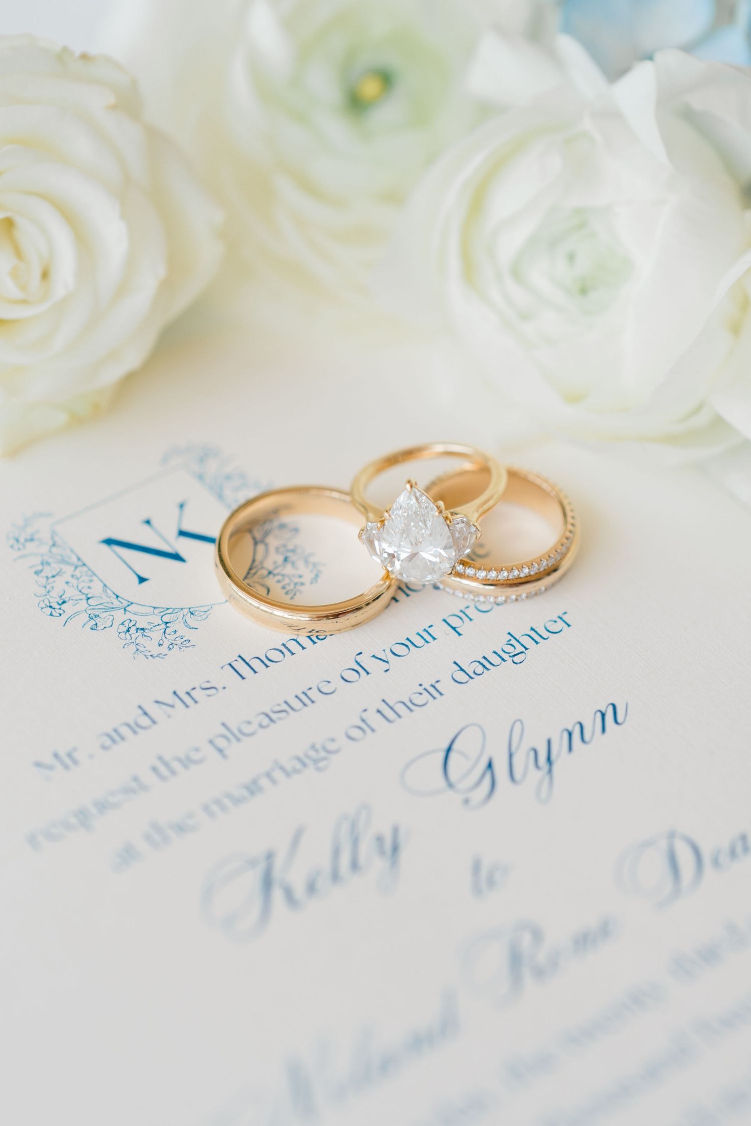 Gold wedding rings and elegant blue calligraphy on wedding invitation card with white floral details.