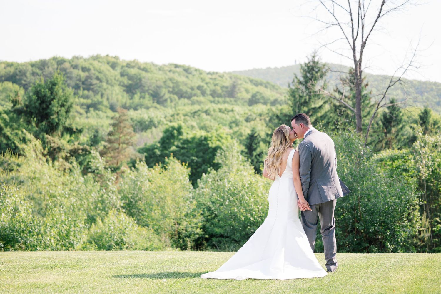 A romantic couple walks together through lush green fields on a sunny summer day while wearing formal wedding attire.