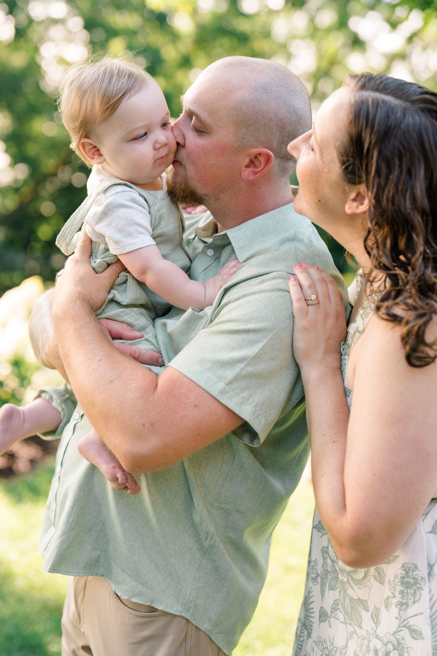 A bald father kisses his baby while a smiling mother in a floral dress looks on outdoors.