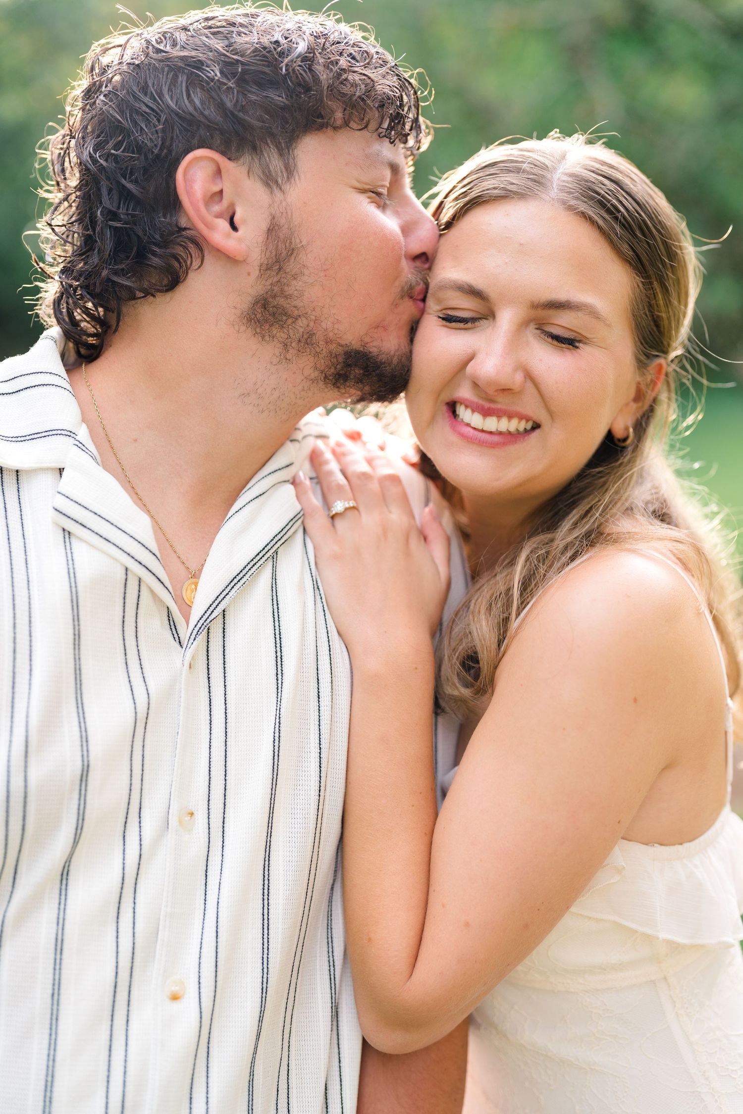 A man kisses a smiling woman on the cheek outdoors, she wears an engagement ring and a white dress.