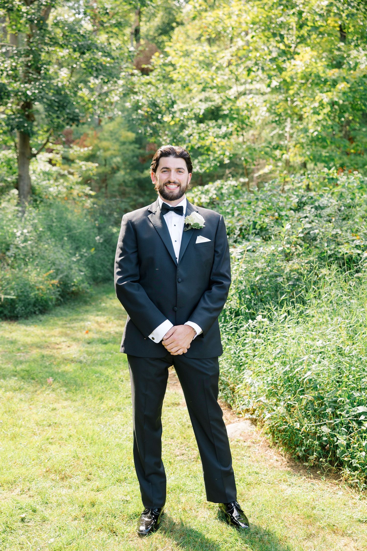 Handsome groom in classic black tuxedo with bow tie and boutonniere smiling outdoors in a green garden setting.