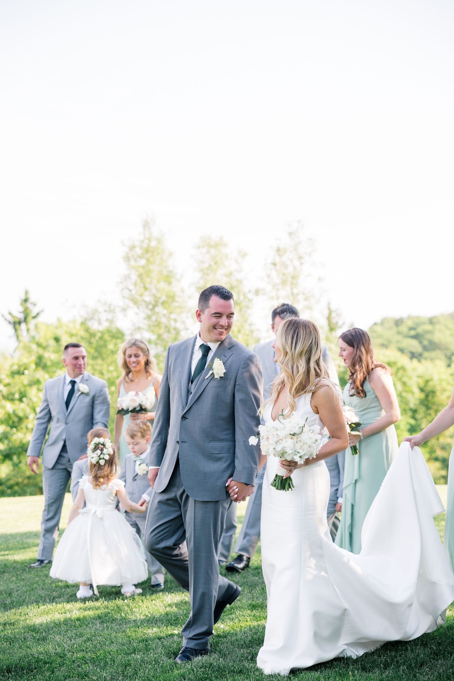 A wedding party walks and laughs together on a grassy hillside on a bright sunny day.
