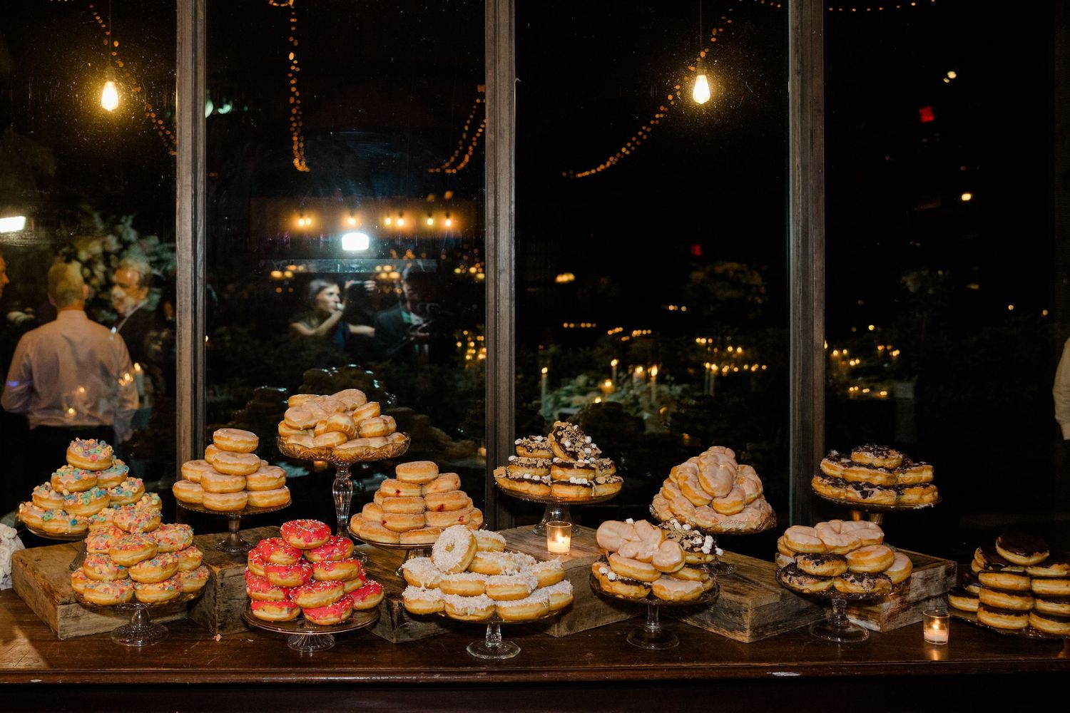 Various tiers of cupcakes and desserts displayed on glass stands against a dark window at night with glowing lights.