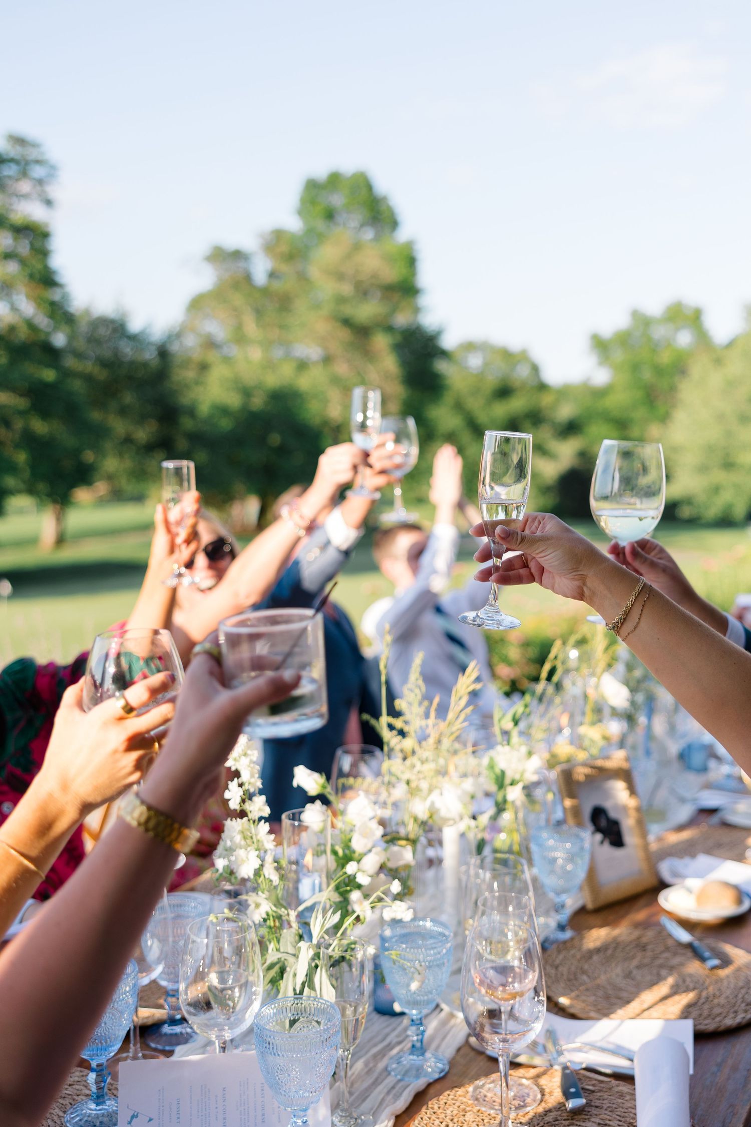 Wedding guests raise champagne glasses in a celebratory toast over a floral-decorated outdoor reception table.