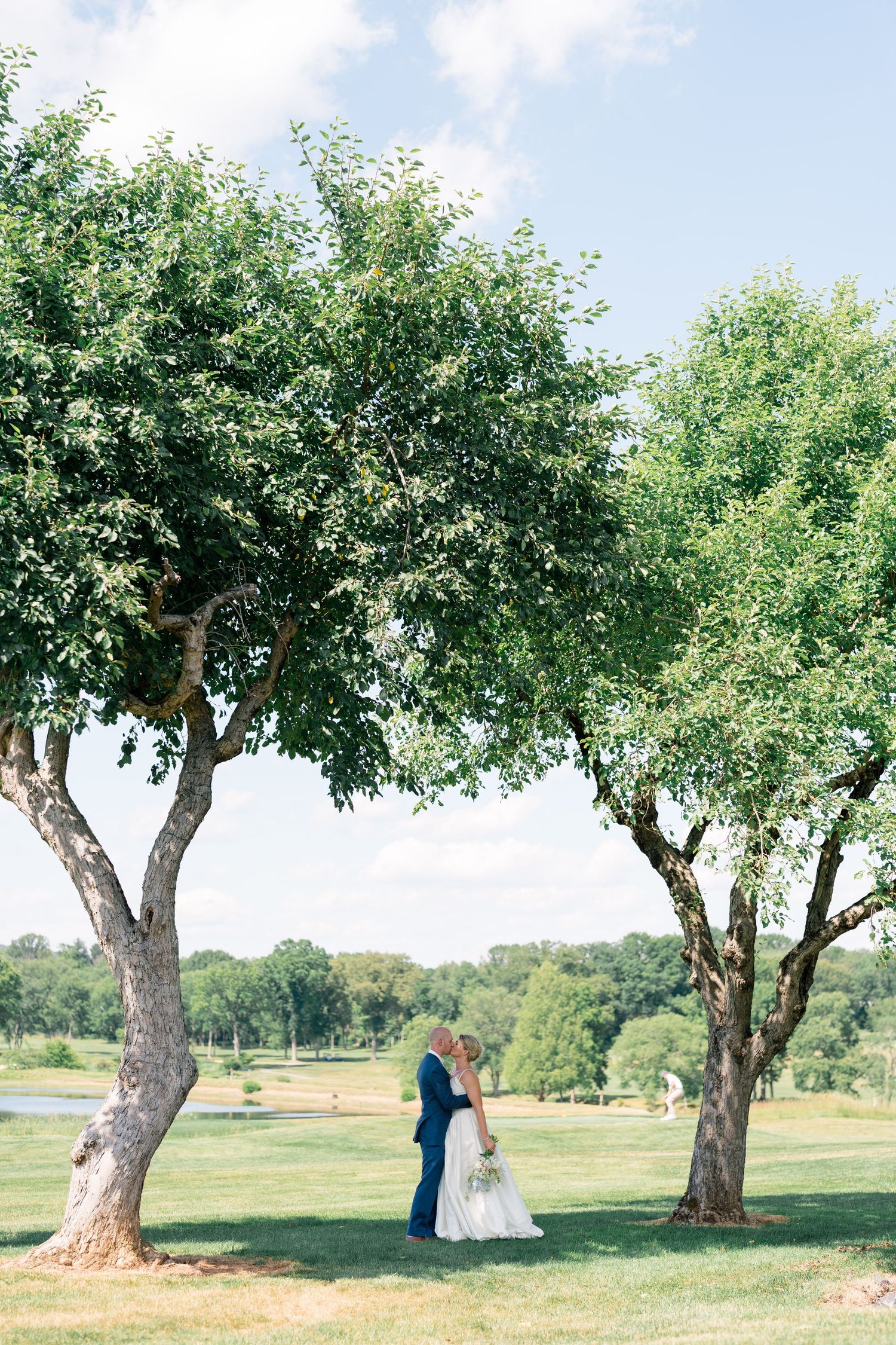Bride and groom share a romantic kiss between two trees on a lush green golf course on their wedding day.