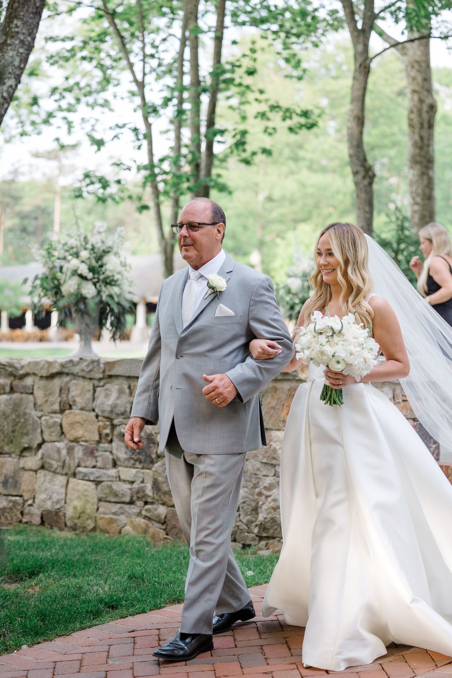 Wedding scene on brick pathway with stone wall backdrop showing couple walking arm in arm during outdoor ceremony.