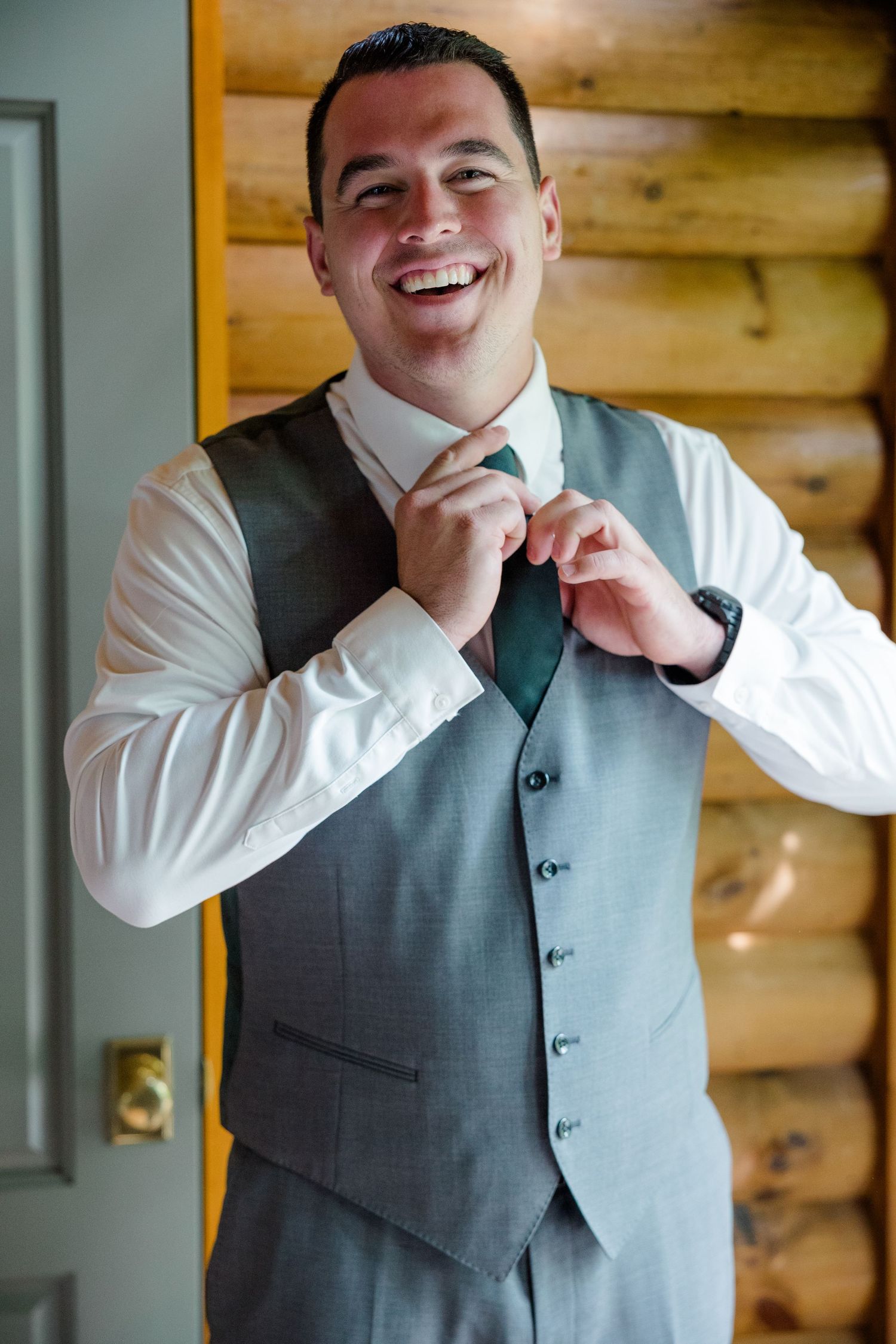 A person in formalwear adjusts a teal tie while wearing a gray vest and white dress shirt inside a rustic log cabin.