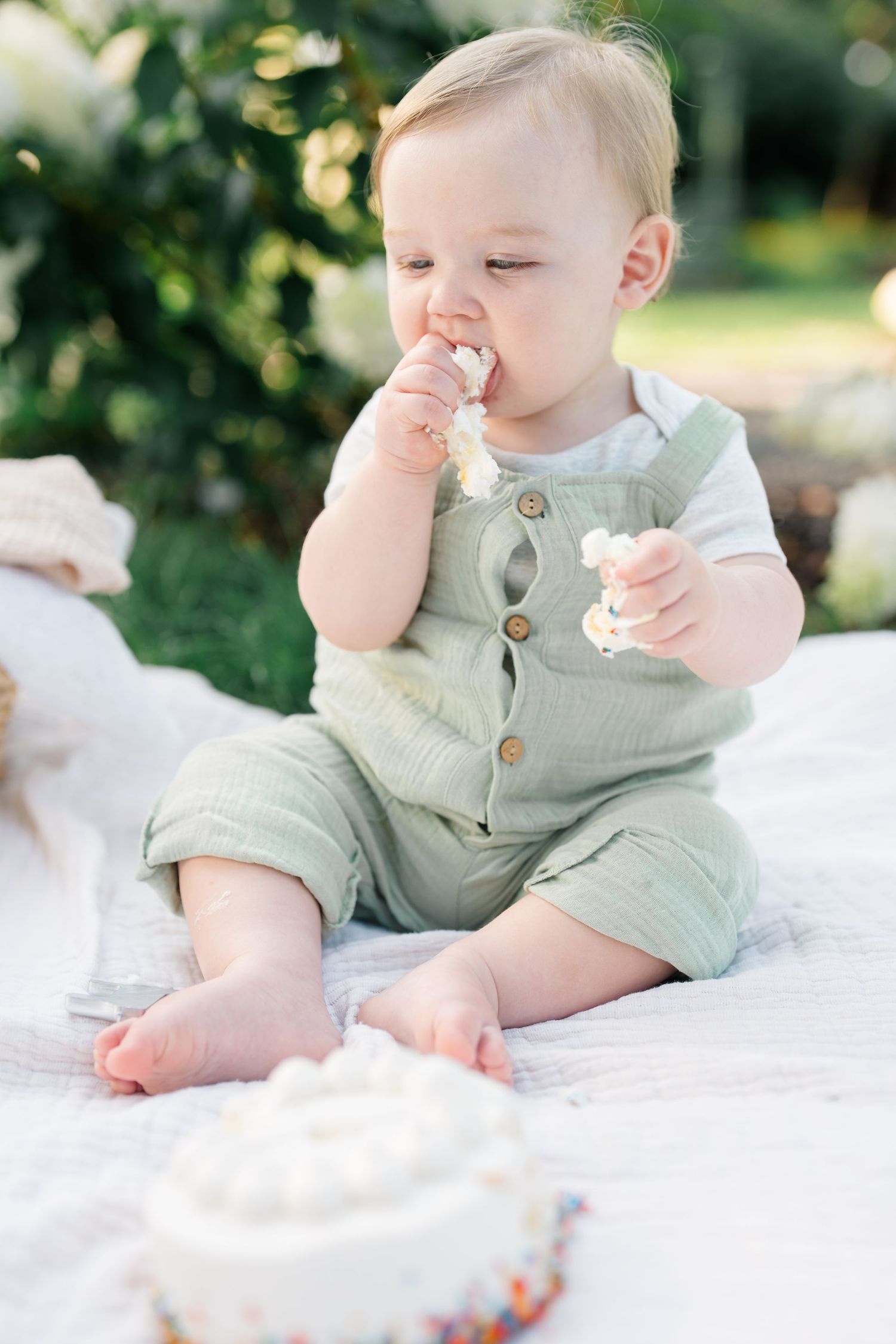 Adorable baby in green overalls enjoys smashing and eating white birthday cake outdoors.