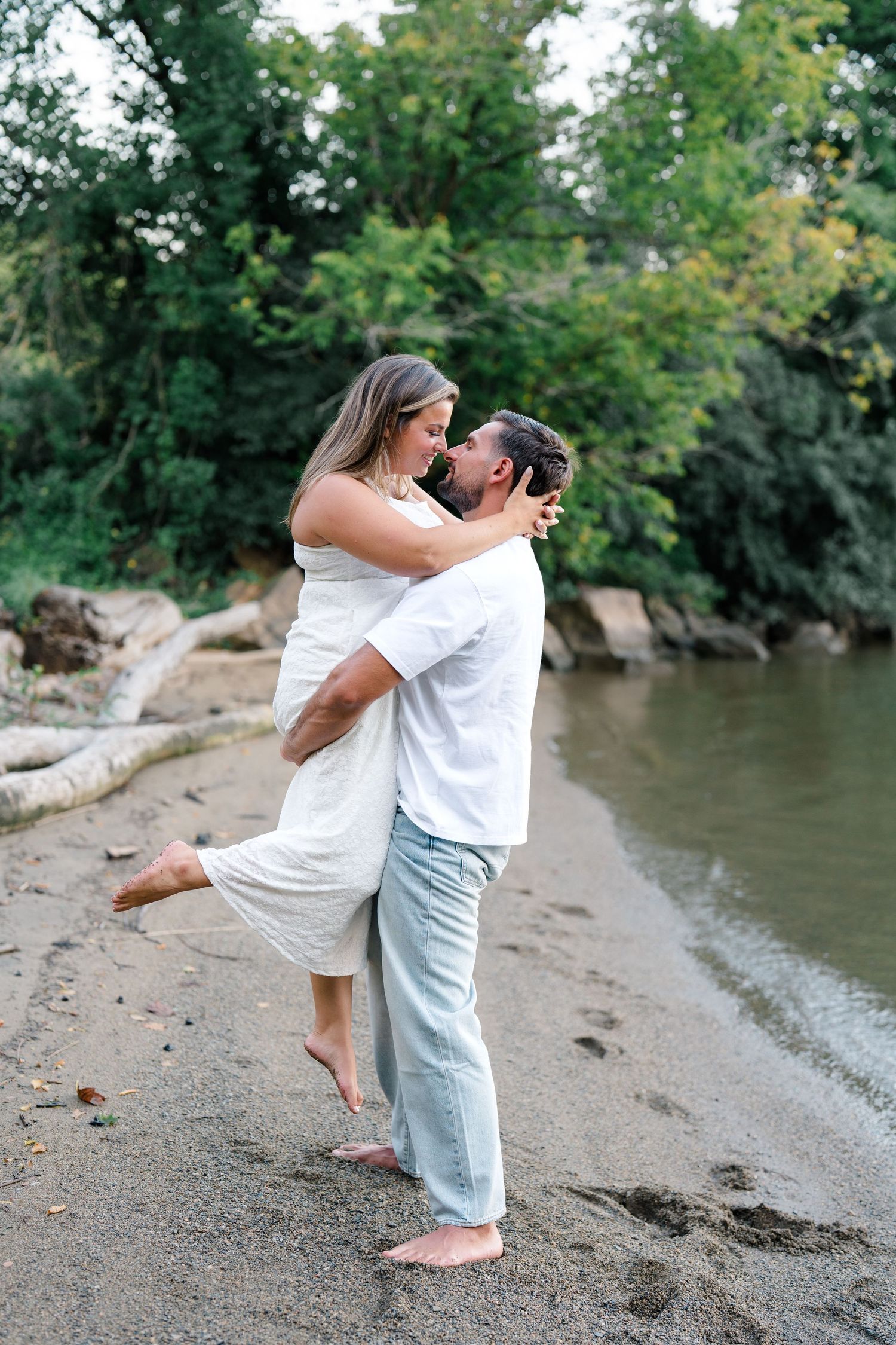 Couple embraces on sandy riverbank, man lifts woman in white dress near lush green trees.