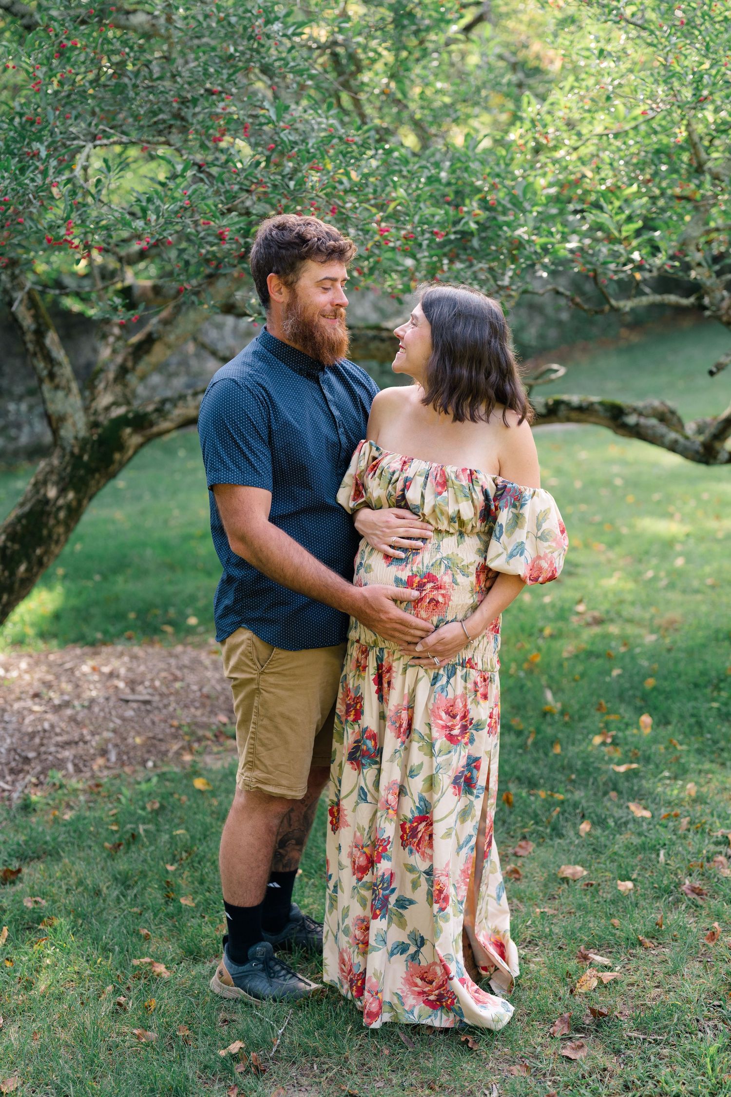 Pregnant couple embracing outdoors under a tree, woman in floral dress, man in blue shirt and khaki shorts.