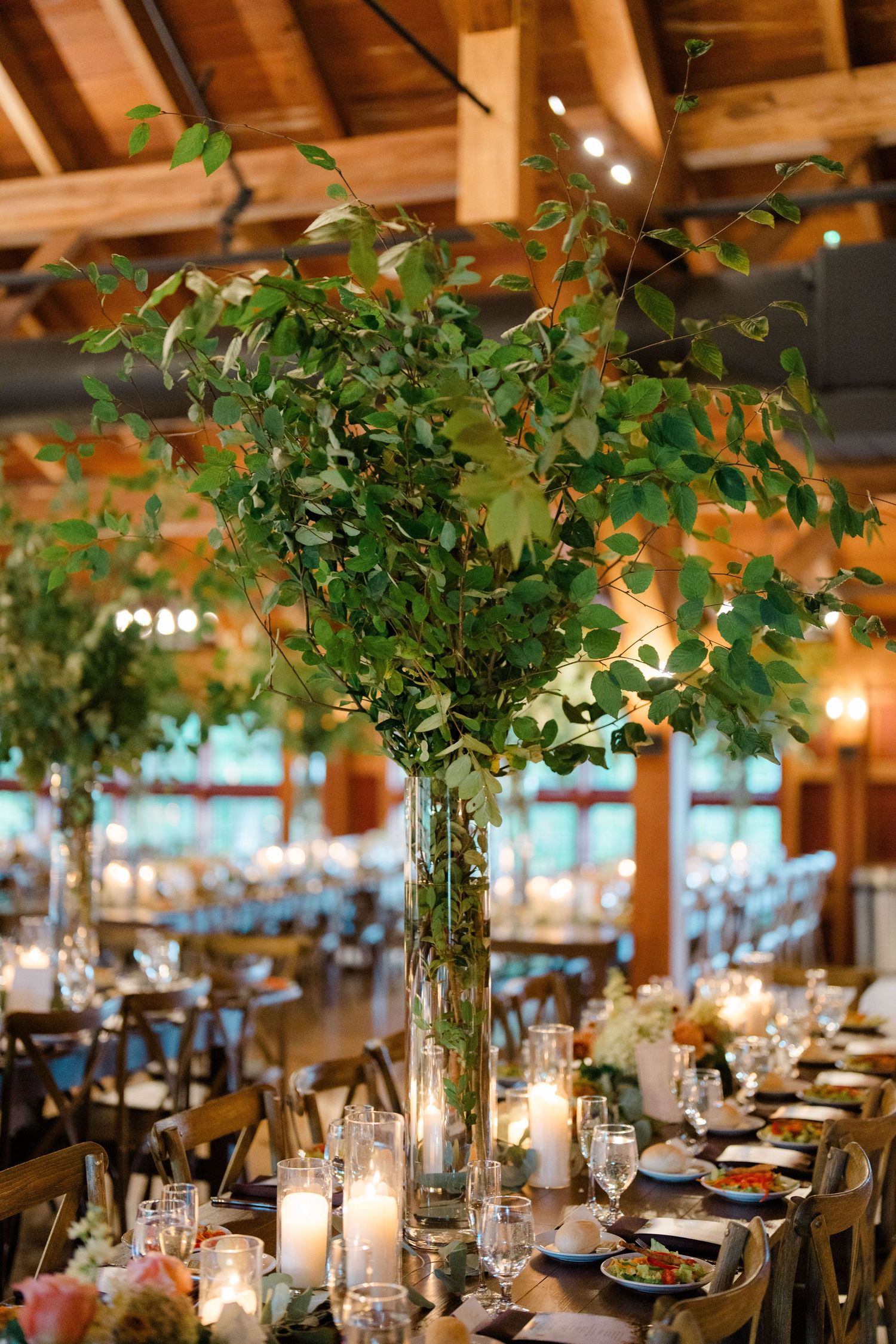 Tall glass vase centerpiece with lush green branches on a candlelit wedding reception table in a rustic wood venue.