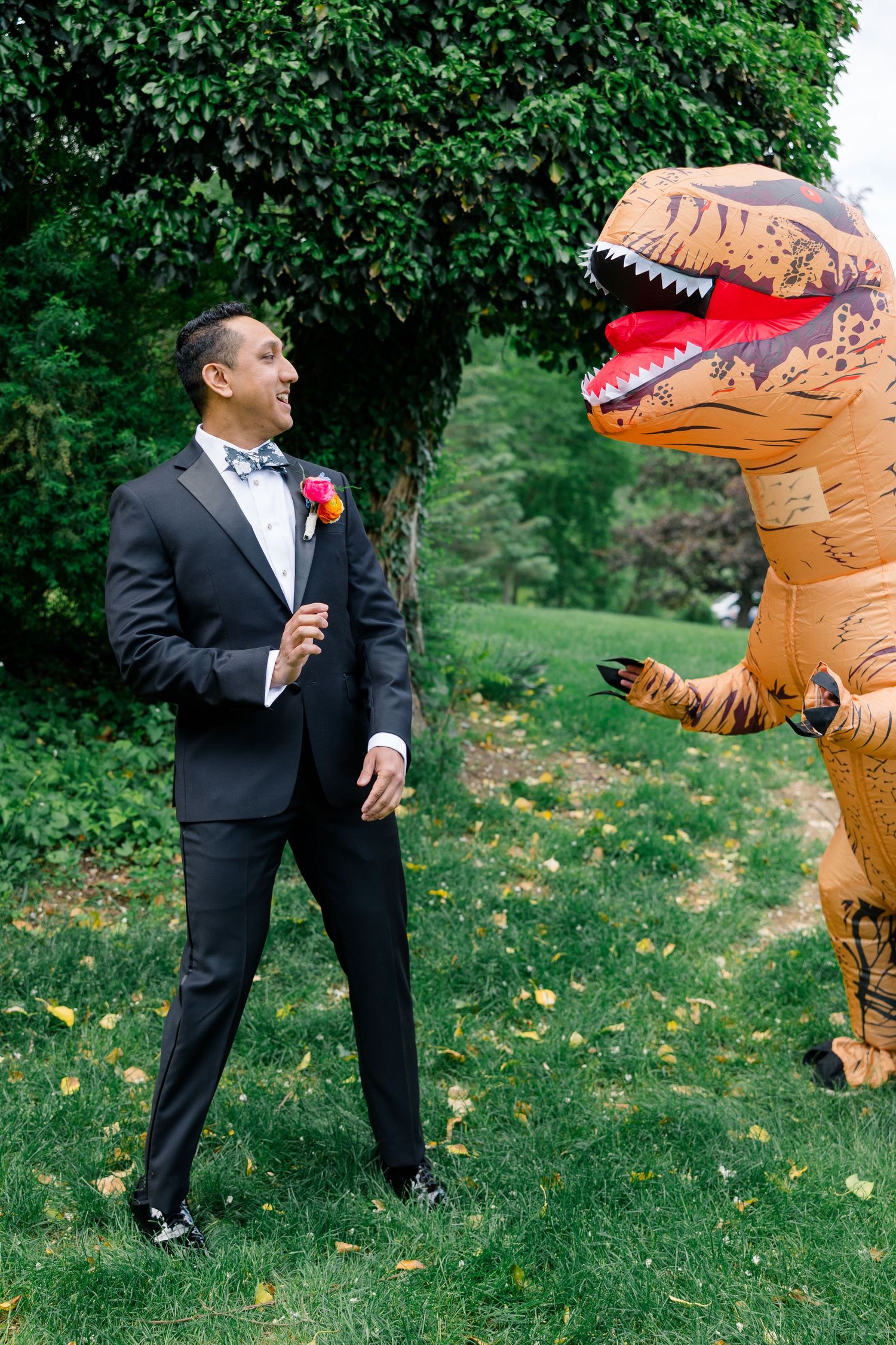 Wedding photo featuring a groom in a tuxedo playfully posing with someone in an inflatable T-Rex costume outdoors.
