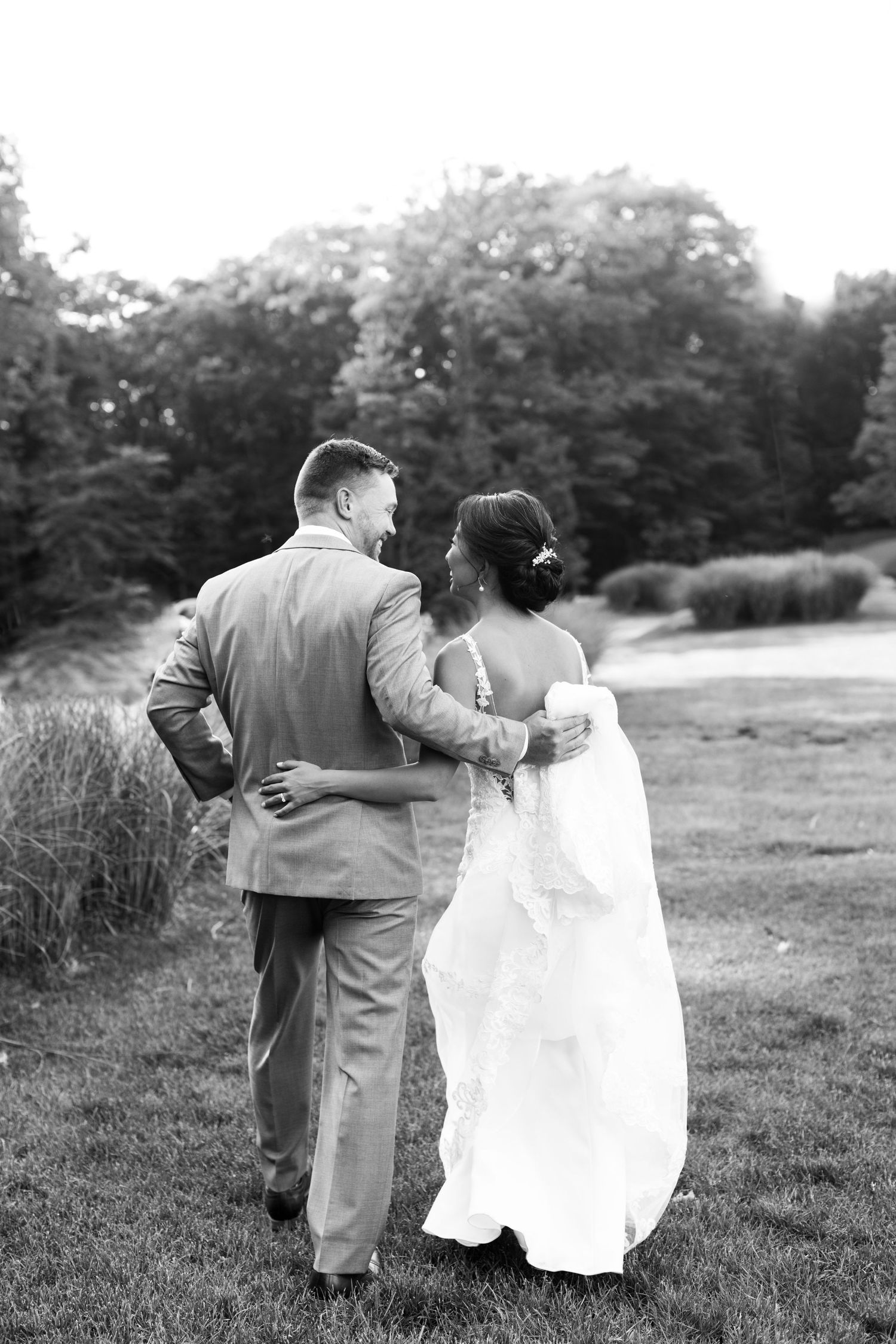 Black and white photo of a couple walking together on grass, with her in a wedding dress and him in a suit.