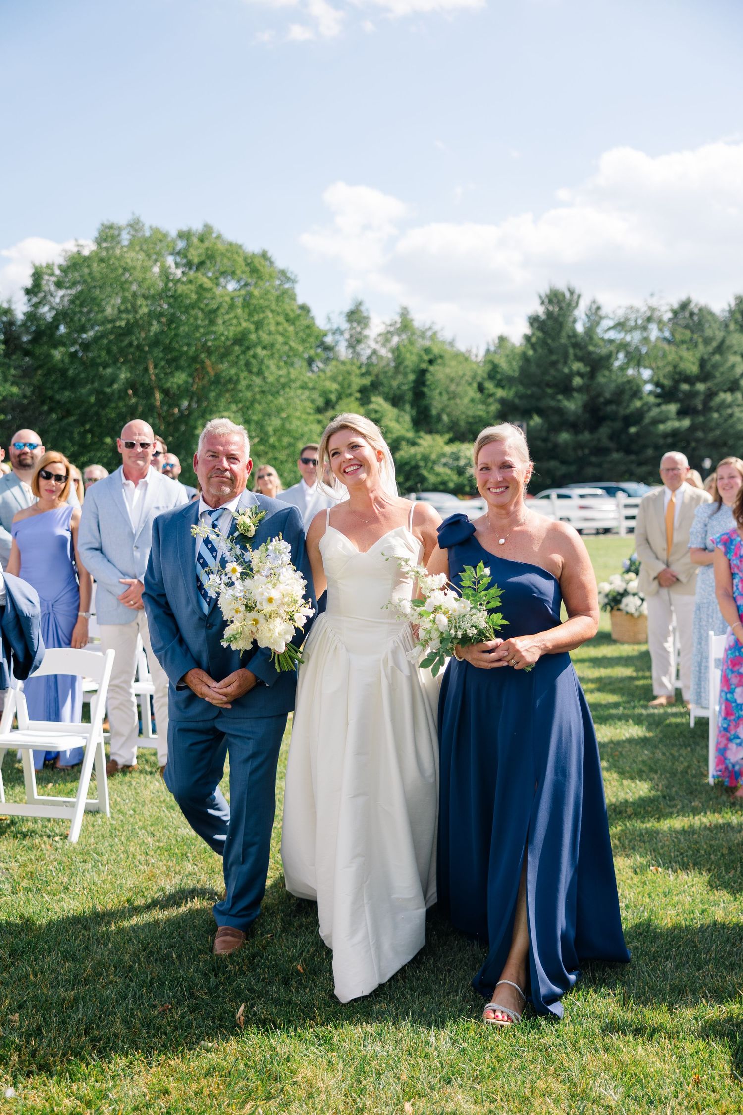 Smiling bride walks down the grassy aisle escorted by two family members at an outdoor wedding ceremony.
