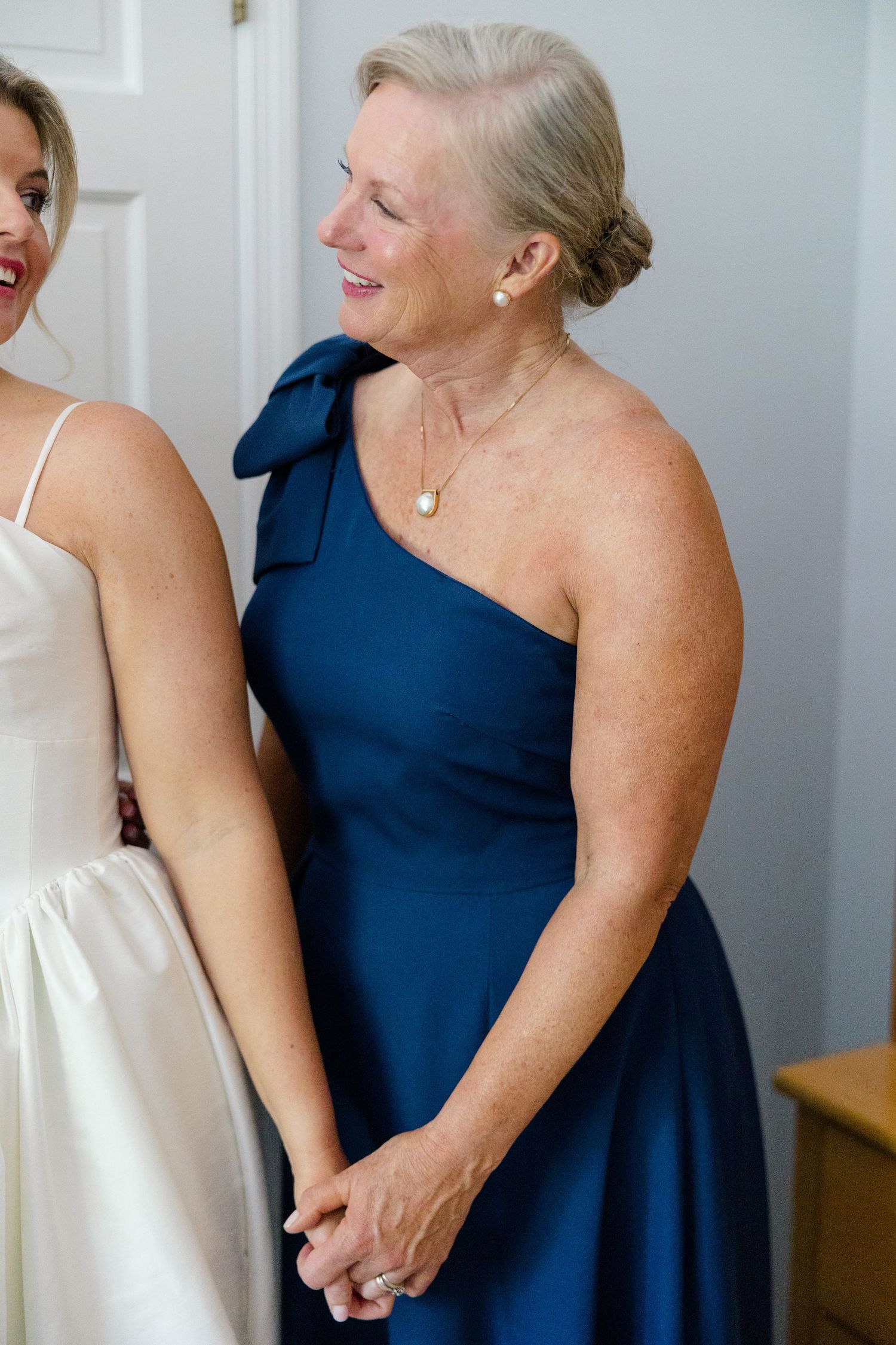 Mother of the bride in navy one-shoulder gown holds hands with bride in white dress during wedding preparations.