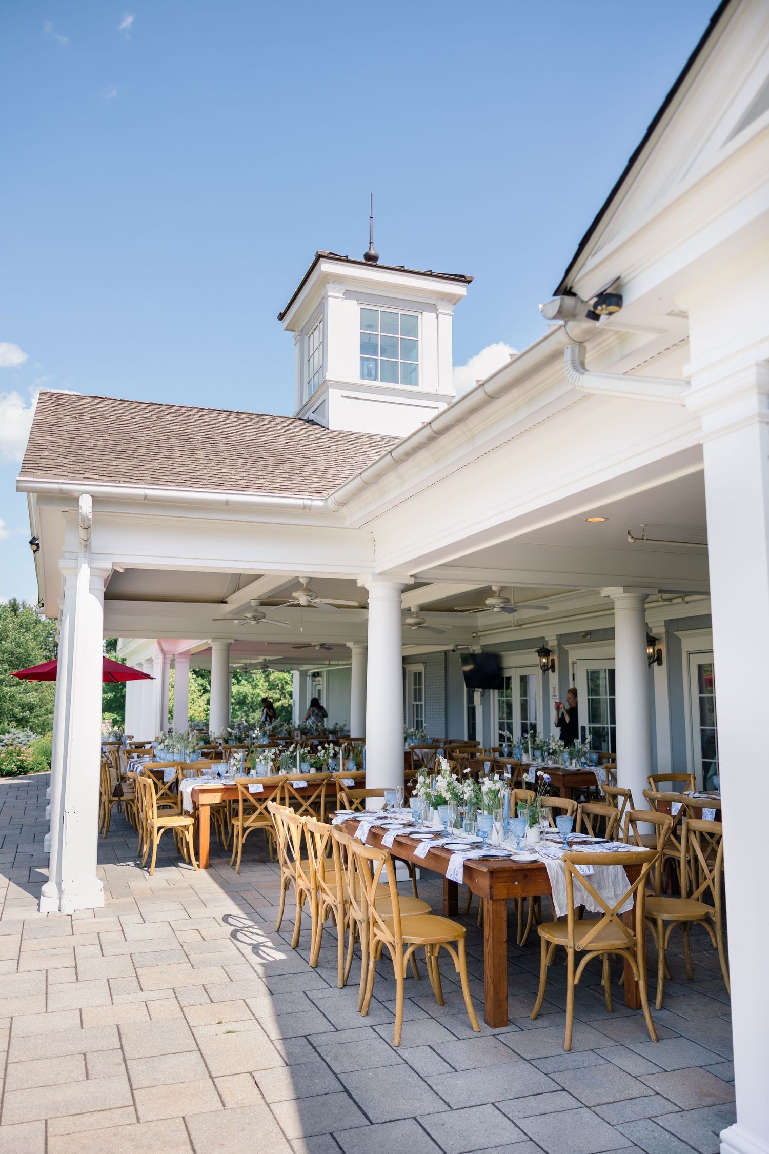 Elegant outdoor wedding reception setup with wooden chairs and long tables under white clubhouse patio.