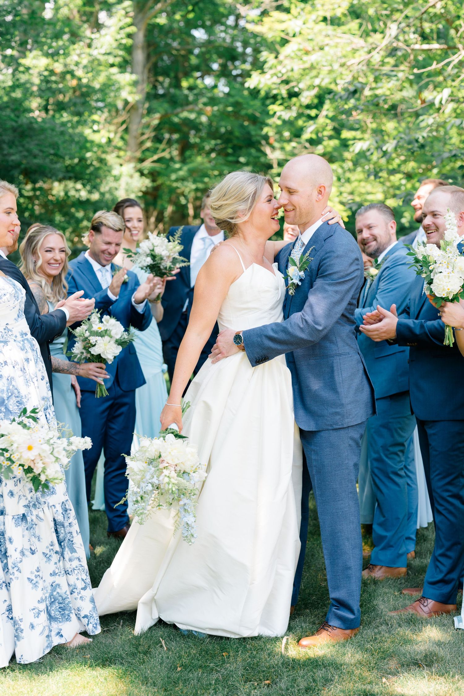 Bride and groom share a joyful kiss surrounded by cheering wedding party guests on a sunny lawn.