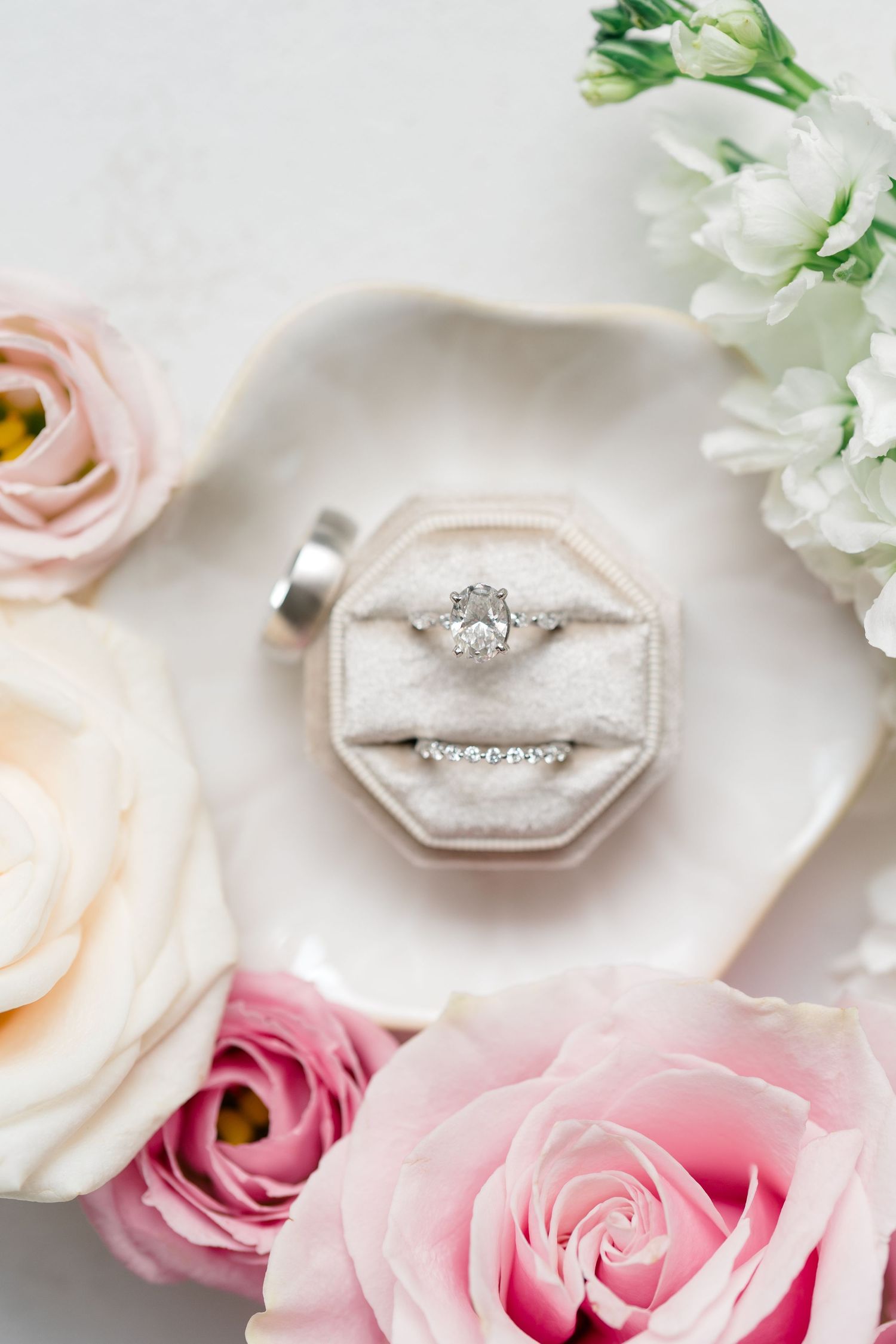 Diamond engagement ring nestled in white velvet box surrounded by pink and white roses.