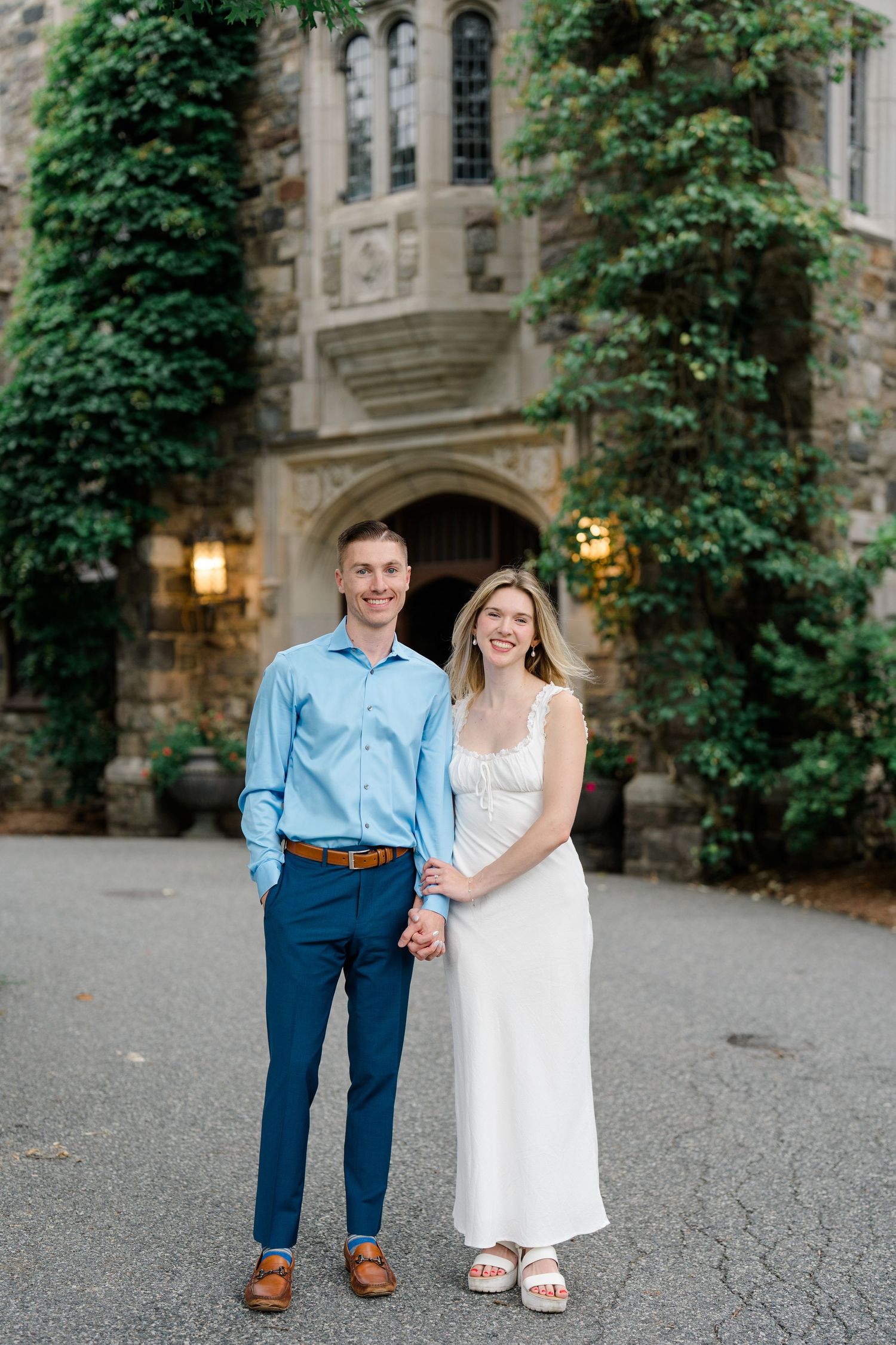 Smiling couple holding hands in front of an ivy-covered stone building, groom in blue suit and bride in white dress.