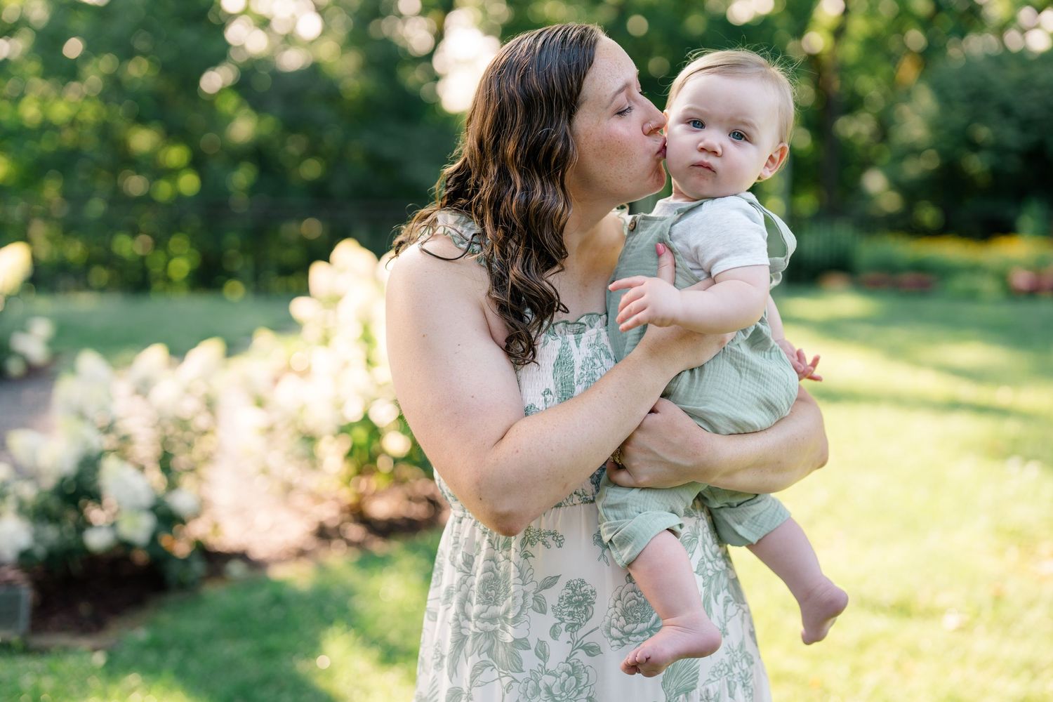 A mother in a floral dress kisses her baby in a green outfit outdoors in a lush garden setting.