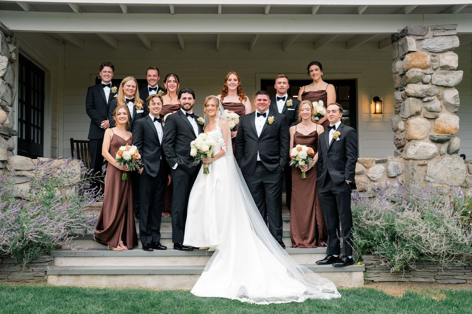 Wedding party poses on stone building steps, bride in white gown with groom in tux, bridesmaids in brown dresses.