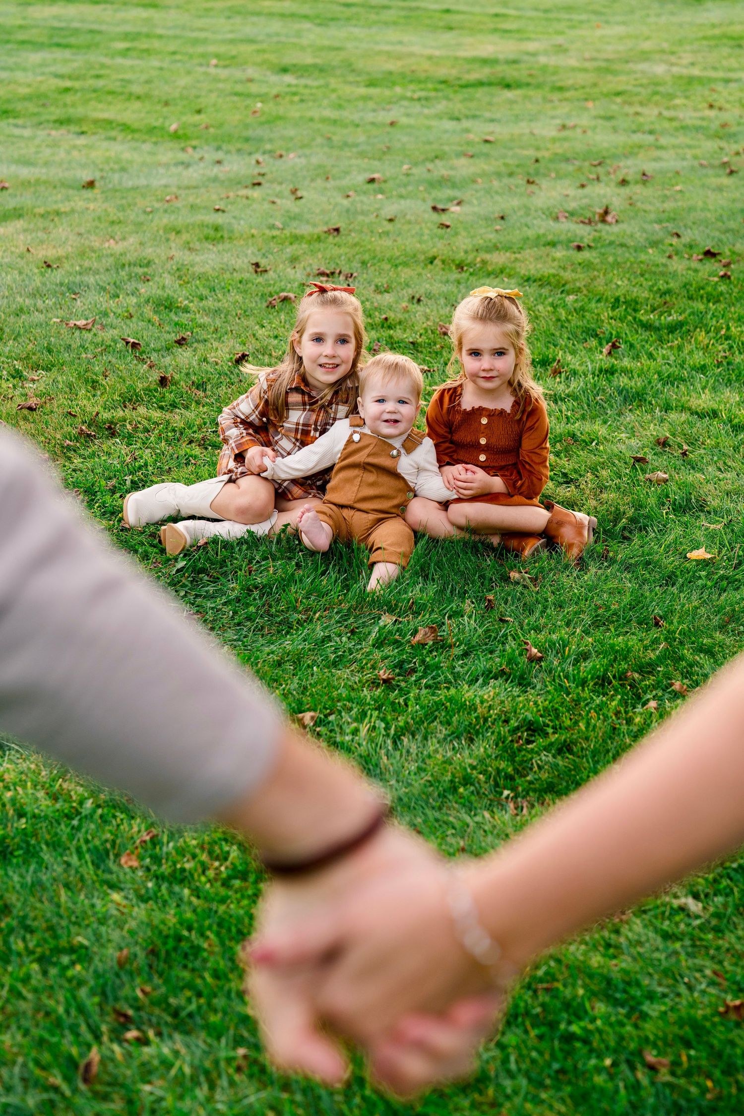 Two adults hold hands while three young children sit smiling on green grass in a heartwarming family outdoor photo.