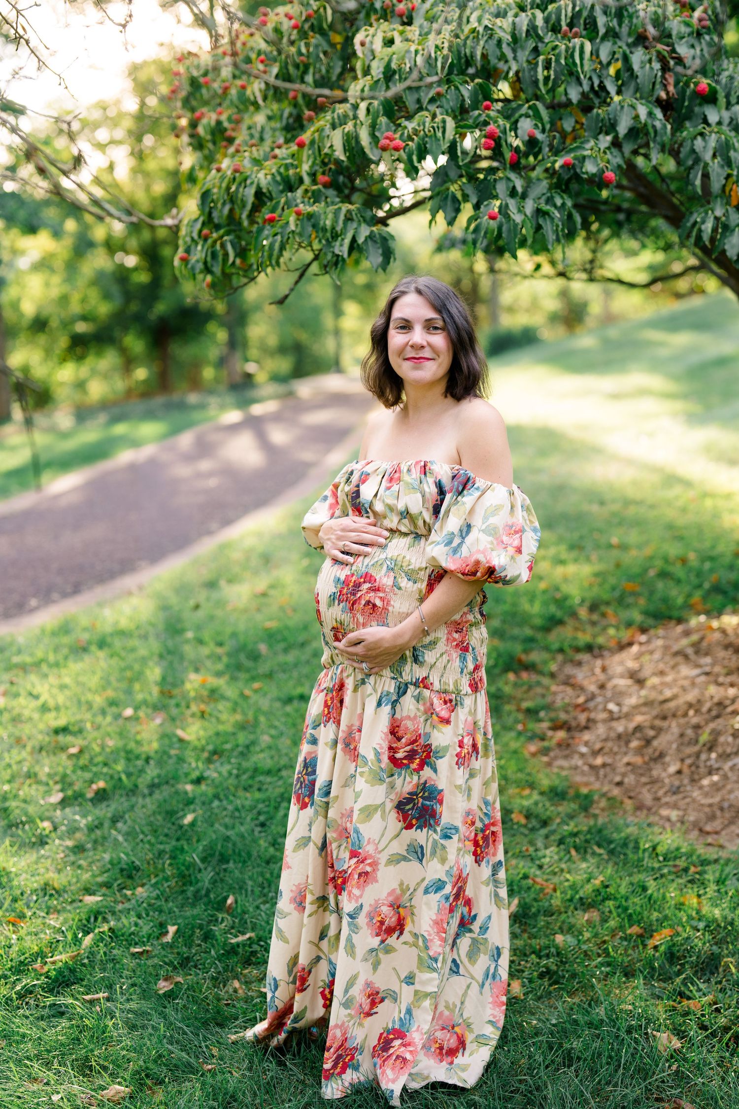 Pregnant woman in floral off-shoulder maxi dress cradling baby bump outdoors in lush green park setting.