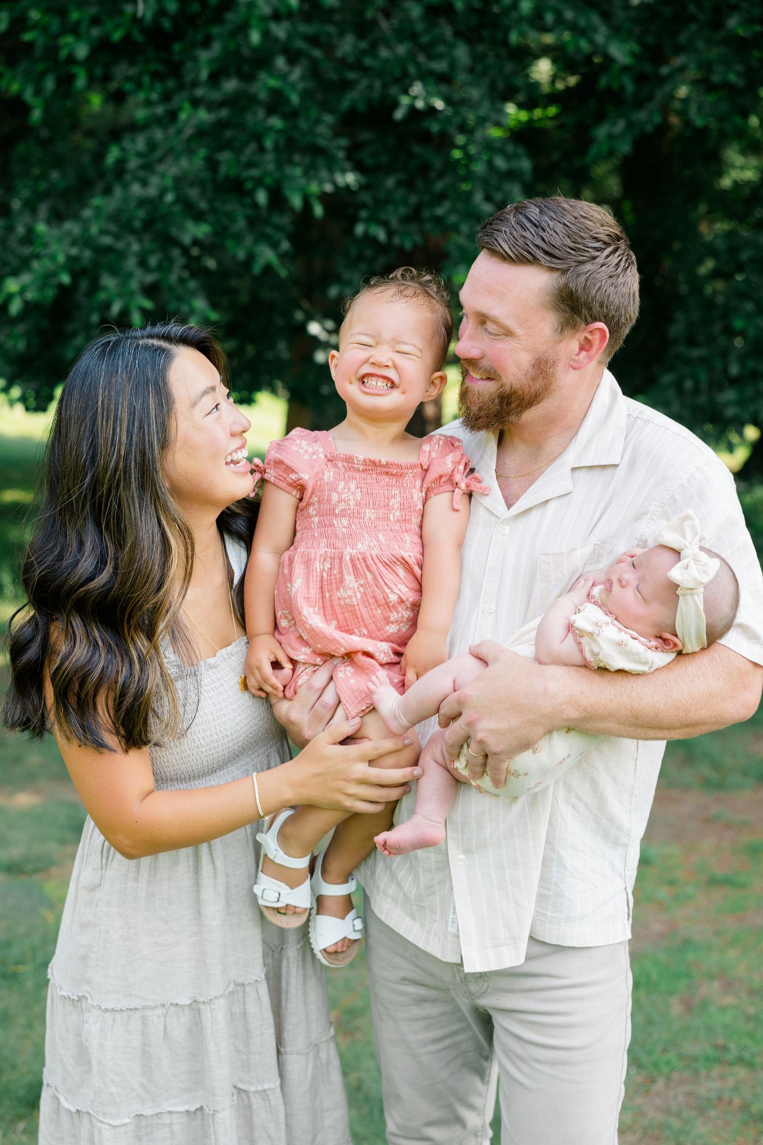 Happy family of four outdoors, parents holding toddler girl in pink dress and newborn baby, smiling together.