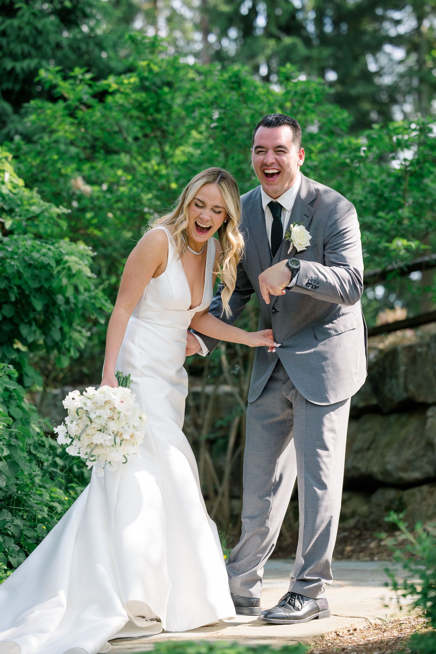A joyful wedding couple laughing together outdoors with the bride holding a white bouquet.